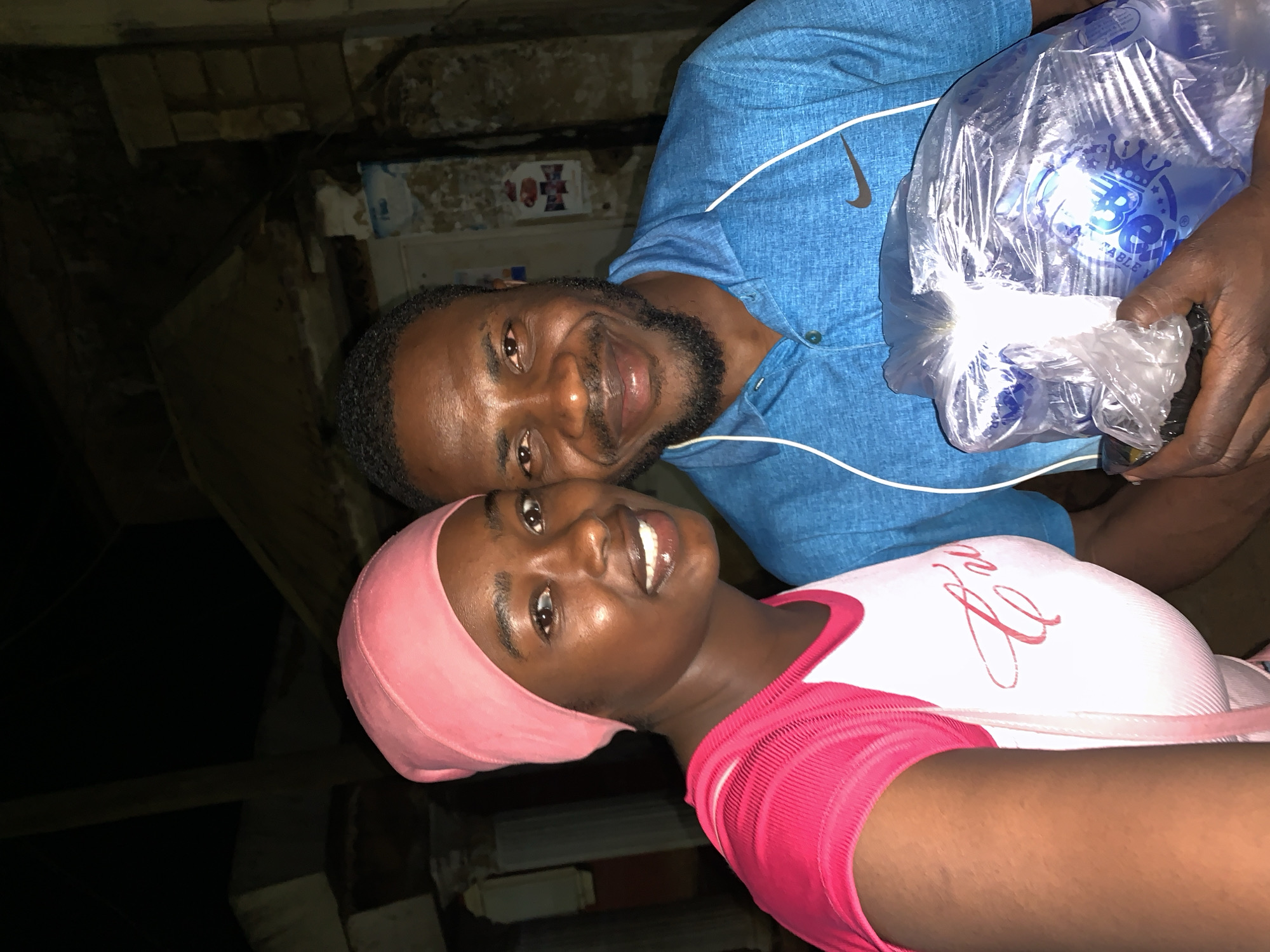 A dark-skinned man and a woman are posing for a photograph in a dimly lit environment, potentially indoors. The man is wearing a blue Nike-branded shirt and is holding a large plastic bag filled with bottled water, labeled with a "B" and a crown. The woman, positioned in front of the man, is wearing a pink head covering and a pink and white top. The background is obscured, but brick and other materials can be discerned. The photograph was taken in Kabong, Nigeria.