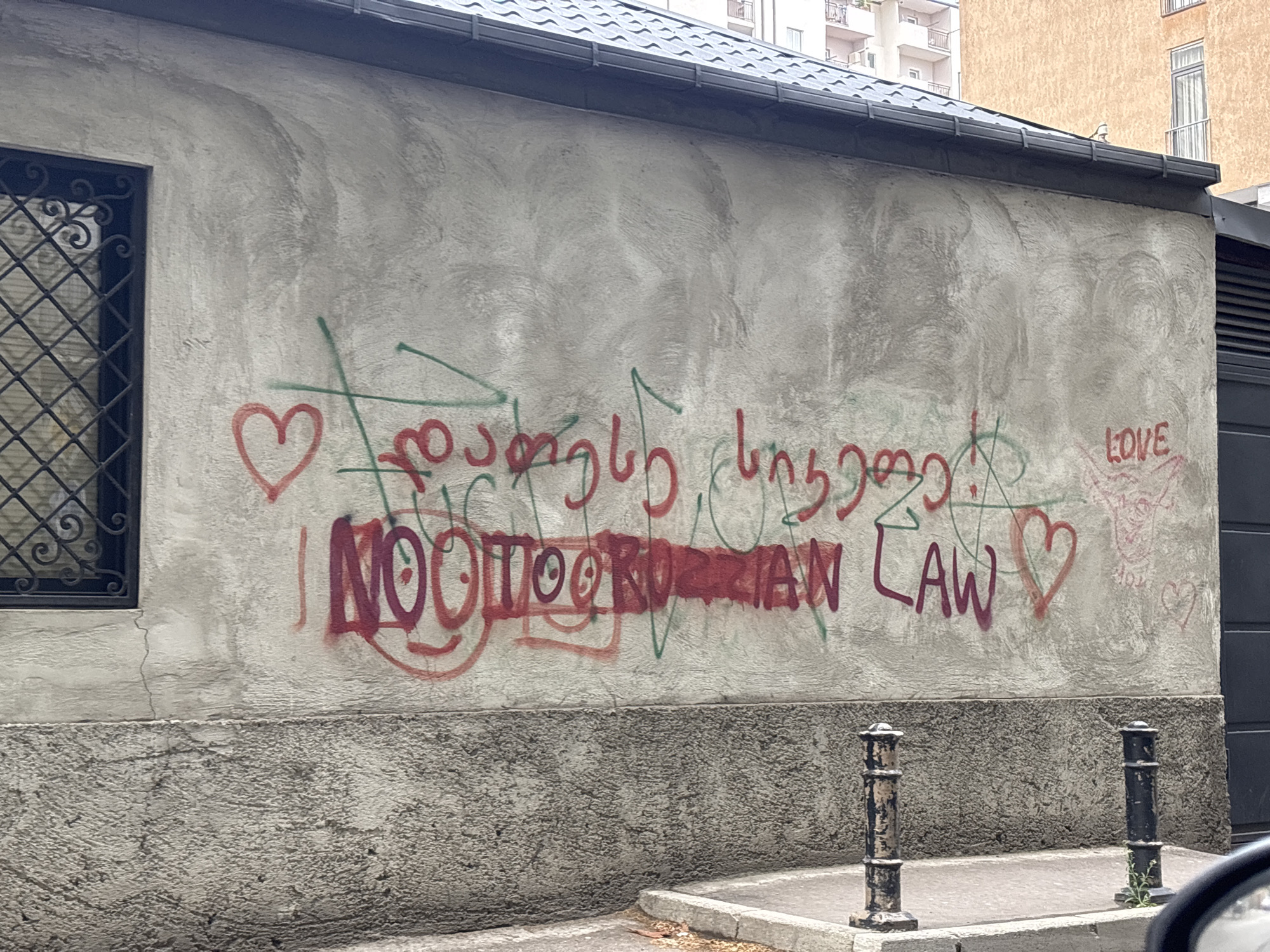 The image displays a weathered concrete wall covered in graffiti, situated in an urban environment, likely Tbilisi, Georgia. The wall, painted a light grey, features a dark metal window with an elaborate grate on the left. To the right, a section of dark, possibly corrugated siding or a gate, is visible. A darker, rough concrete base runs along the bottom of the wall, with two black and bronze-colored bollards standing on a sidewalk in front. Above the wall, a dark tiled roofline is present, and in the background, apartment buildings with balconies can be seen under an overcast sky.
The graffiti is a prominent feature, rendered in red, green, and purple spray paint. In the center, the Georgian word "თავისუფლება" (tavisupleba), meaning "freedom," is written in green and red. Below this, a bold English message in red and purple reads "NOO" with the 'O's stylized as smiley faces, followed by a stylized word that visually resembles "TOOR" (also with smiley faces for 'O's) or a stylized "FOR", and then "RUSSIAN LAW". This is a common protest slogan against the "foreign agent law". Several red hearts are scattered across the wall, and the word "LOVE" is written in pink/red in the upper right corner. The scene depicts a daytime setting with diffused light.
