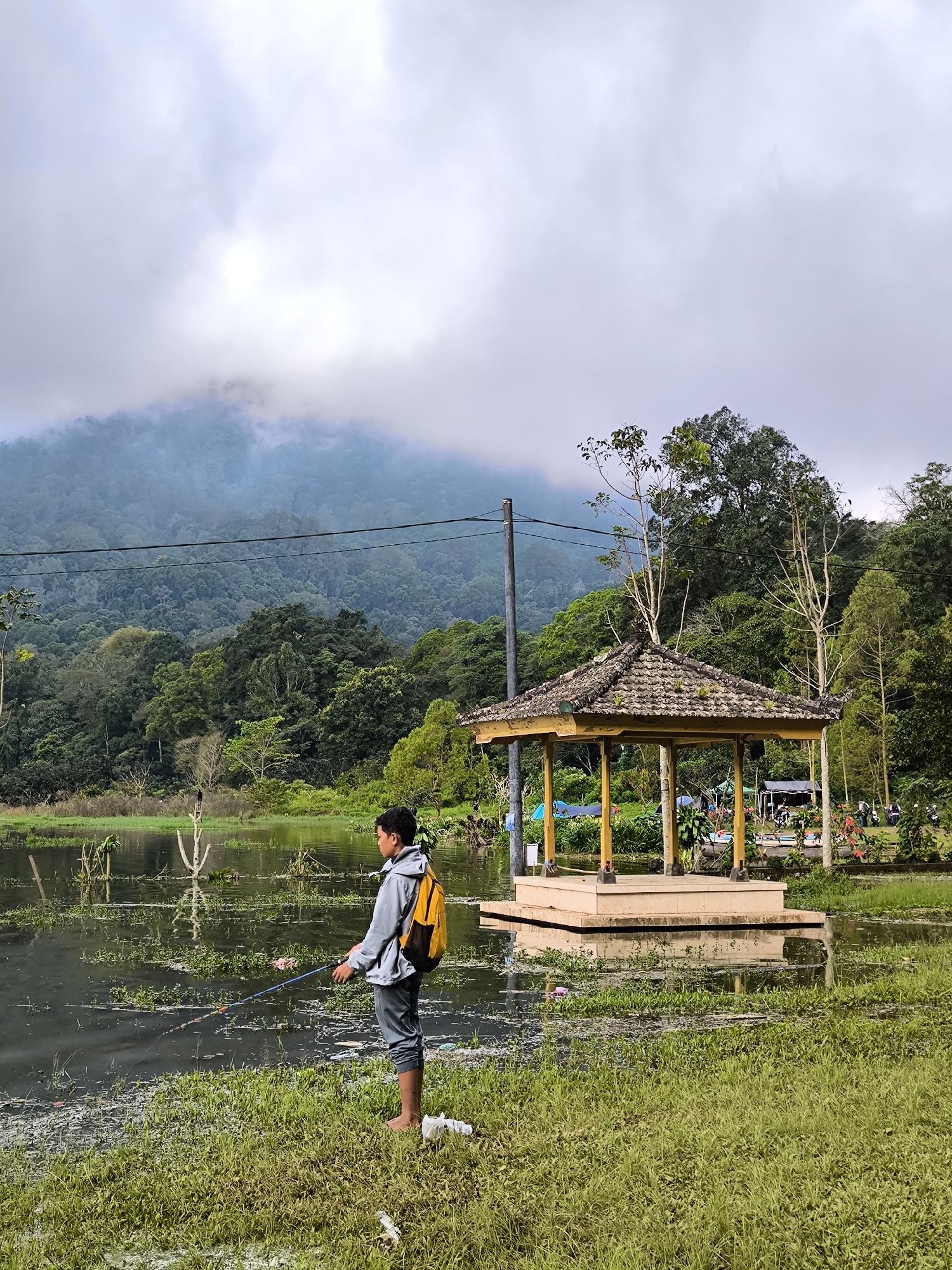 A person is fishing in a flooded area near a gazebo. The individual is standing barefoot in grassy vegetation, wearing a gray jacket and pants, with a yellow backpack. They are holding a fishing rod. The water level appears high, flooding up to the base of the gazebo. In the background, there is a heavily wooded hillside partially obscured by fog or clouds. An electrical pole is visible on the left, while tents and other people can be seen further back near the gazebo. The location appears to be a recreational area in Unknown, Indonesia.