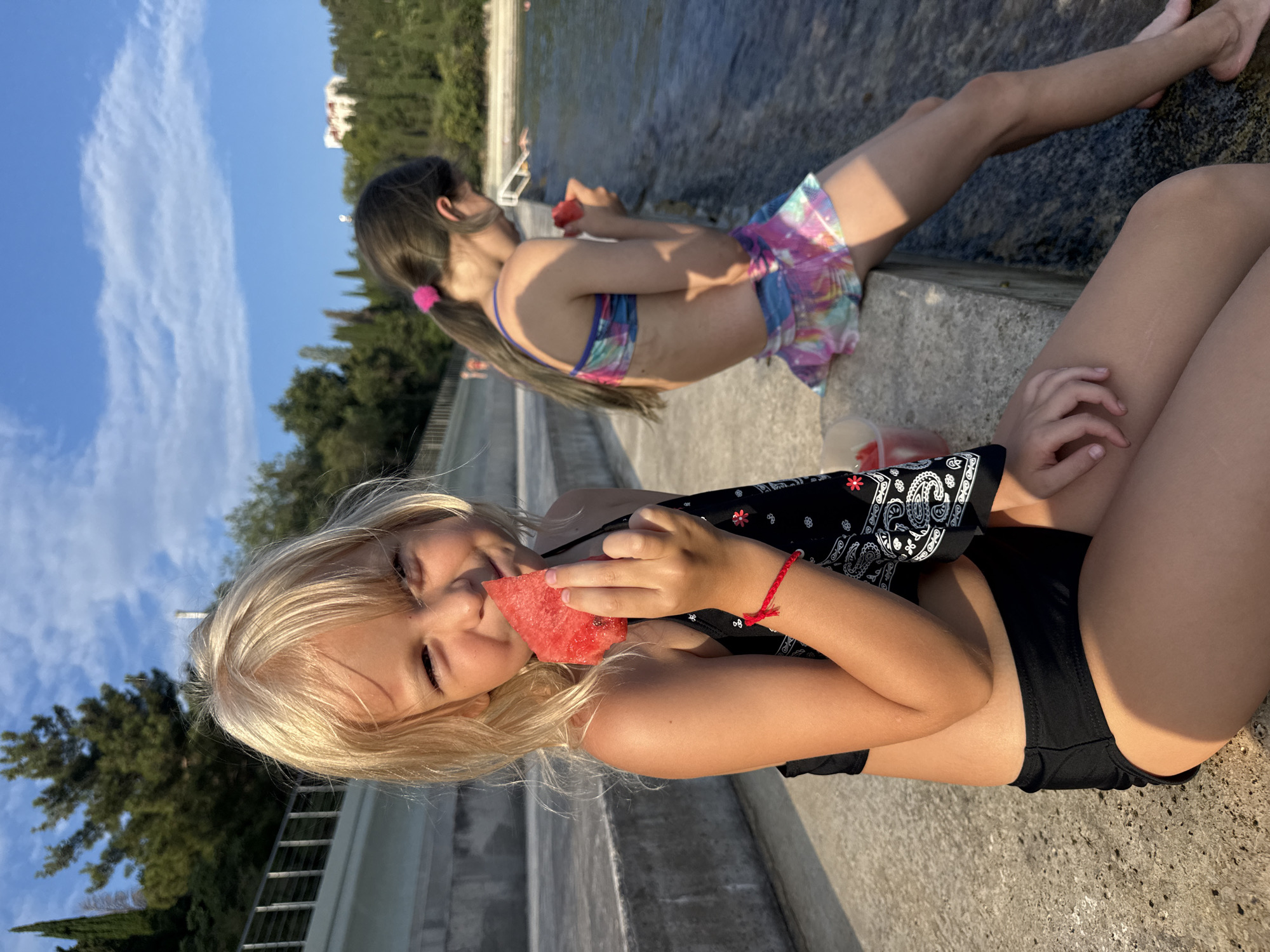 In this media file, two young girls are seen outdoors on a sunny day, likely during warm weather. The scene appears to be a public outdoor area, possibly a park or recreational facility, with concrete structures and lush green trees in the background under a bright blue sky with scattered clouds.
The girl in the foreground, with blonde hair, is wearing a black bikini top and bottoms, and is in the process of eating a slice of watermelon. She is looking downwards, and her expression suggests enjoyment. She has a red bracelet on her wrist. A black bandana with a white paisley pattern is visible on her person.
The second girl, positioned further back, has dark hair tied in pigtails with pink elastics. She is wearing a colorful bikini with a tie-dye pattern, paired with a matching skirt. She is also holding a piece of watermelon, and appears to be smiling.
The overall atmosphere is relaxed and cheerful, indicative of a pleasant summer day spent enjoying snacks outdoors. The sunlight casts a warm glow on the scene, and the clear sky suggests good weather. The image has been rotated 90 degrees clockwise. No visible text is present in the media file.