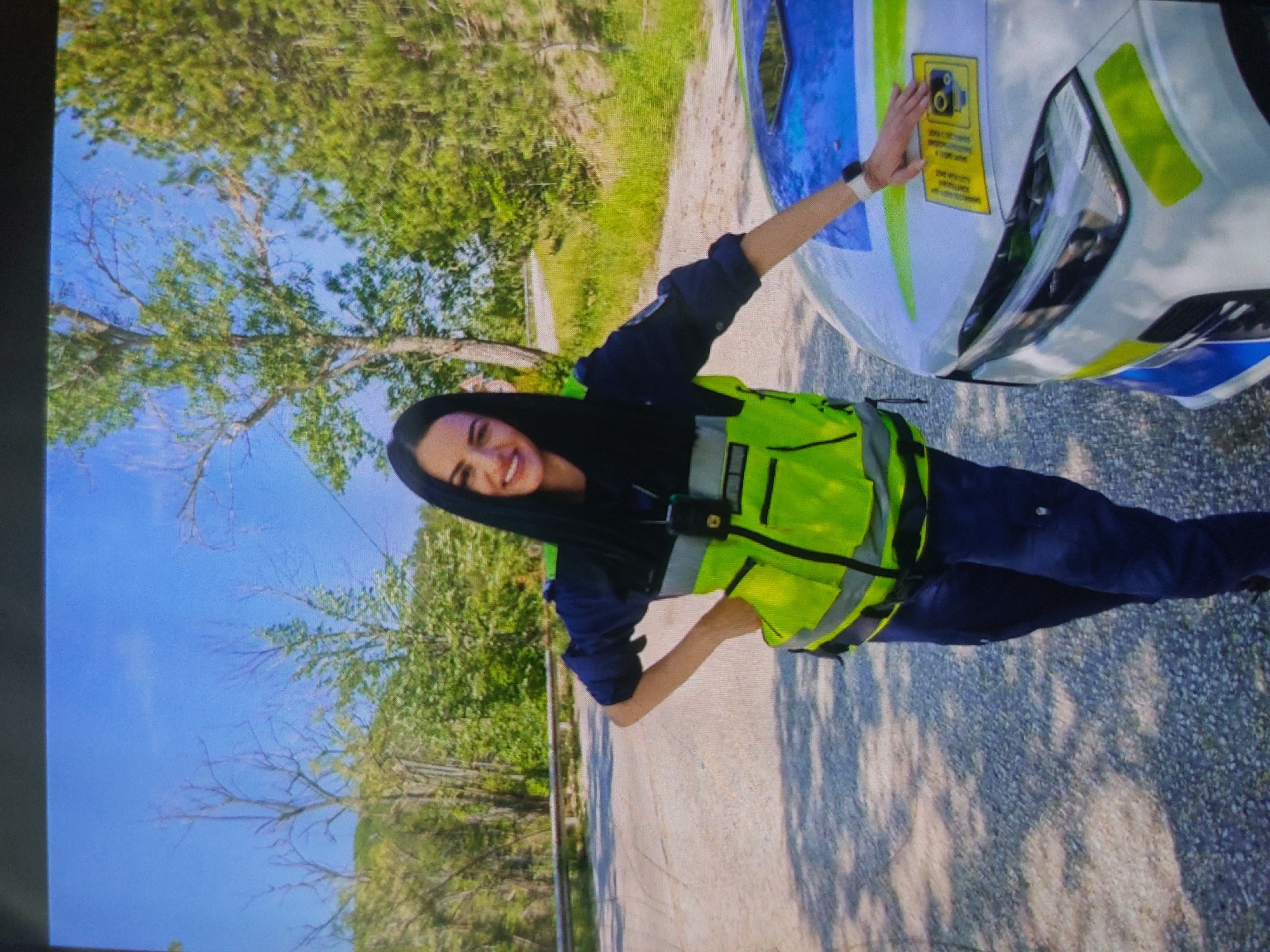 A medium shot depicts a woman in a dark blue uniform and a neon yellow safety vest standing on a gray asphalt road next to a white police car with blue and yellow markings. The woman is smiling and has her arm outstretched, touching a yellow sign on the hood of the police car. Green trees and a blue sky are visible in the background. The location cannot be identified as Tsarevo, Bulgaria from the image alone.