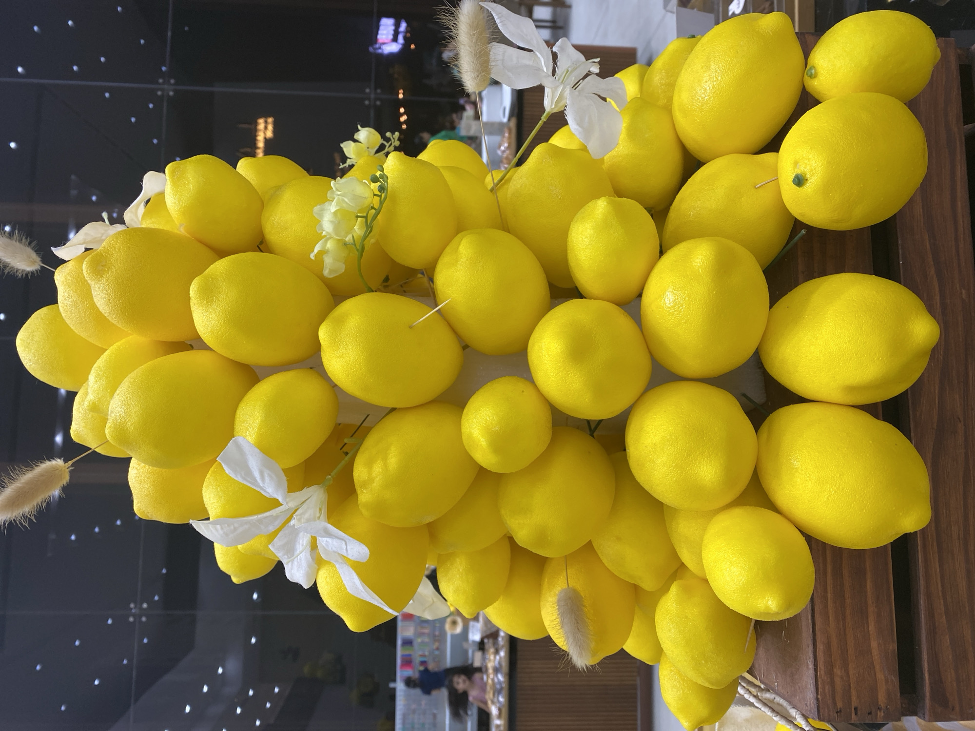 An artful display of bright yellow lemons is arranged in a tiered pyramid shape. The lemons are interspersed with delicate white artificial flowers and fluffy, dried ornamental grasses. The arrangement sits on top of a dark wooden crate. In the blurred background, a glass partition reveals a retail environment. A young girl in a pink shirt is visible in the middle ground, looking towards the lemons. The lighting suggests an indoor setting, possibly a hotel lobby, a restaurant, or a market in Đà Nẵng, Vietnam, given the context provided.