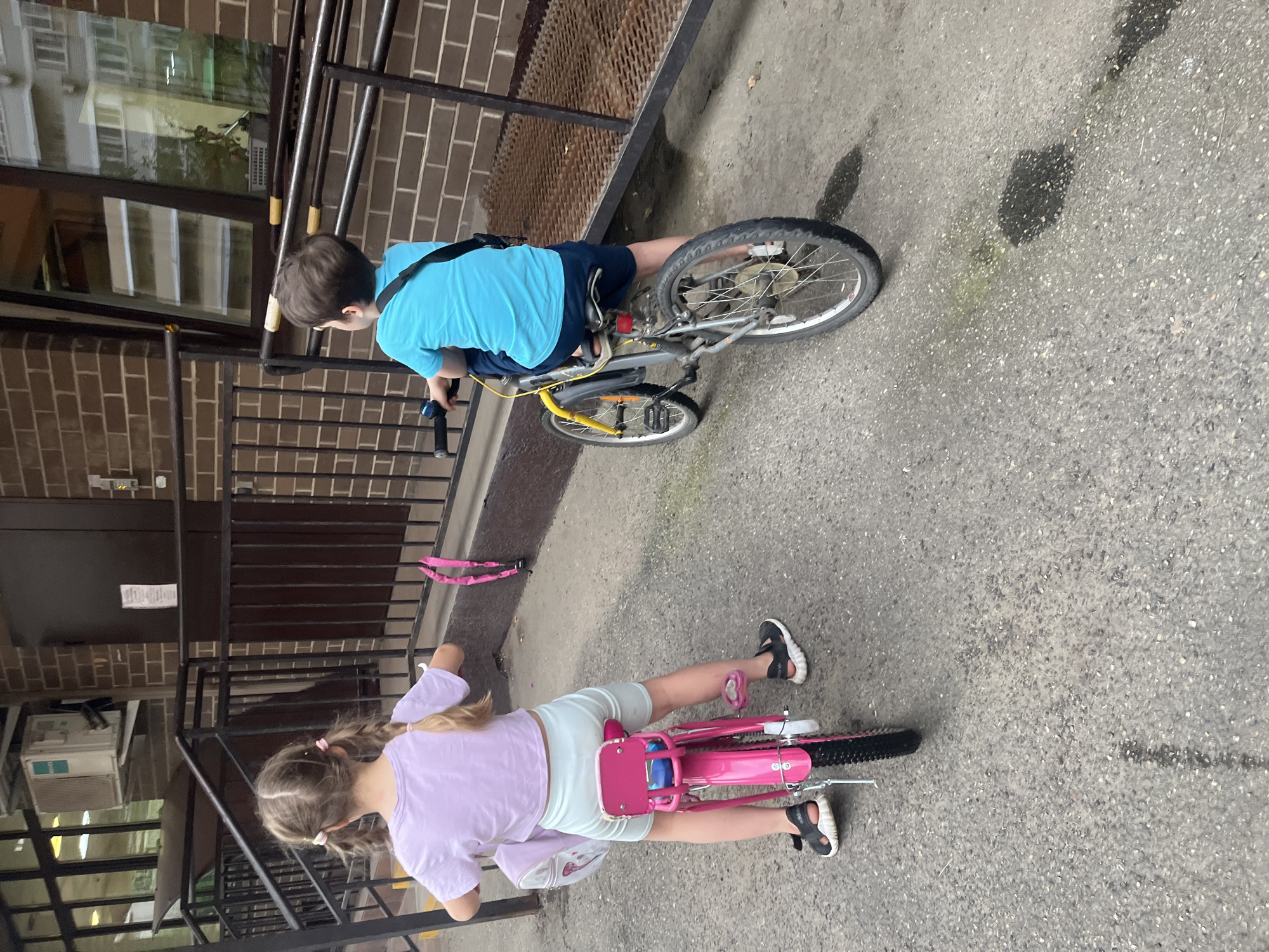 This image captures an outdoor scene in Krasnodar, Russia, depicting two children on bicycles in front of a brick building.
**People:**
1. **Left Child (Girl):** A young girl, viewed from the back, with light brown hair styled in two pigtails secured with pink ties. She wears a light purple short-sleeved crop top, light blue shorts, and black sandals with white soles. She is positioned on a pink bicycle, with a light-colored backpack visible on her back. Her left foot is on the ground, and a kickstand is extended from her bicycle.
2. **Right Child (Boy):** A young boy, viewed from the back, with short dark brown hair. He wears a bright blue short-sleeved t-shirt and dark blue shorts. A black strap crosses his back diagonally, possibly from a small bag. He is seated on a grey bicycle with yellow front forks. His head is turned slightly to his right.
**Objects and Environment:**
* **Bicycles:** The girl's bicycle is predominantly pink with black tires and a silver handlebar. The boy's bicycle is grey with yellow front forks, black handlebars, and black tires, featuring orange wheel reflectors.
* **Building:** A multi-story building constructed with brown brickwork serves as the background. It features multiple dark-framed windows reflecting the surroundings. An external white air conditioning unit, branded "NOVA," is mounted on the brick wall above a window on the left side of the building.
* **Railing and Ramp:** A dark brown metal railing system runs horizontally across the mid-ground. On the right side, a metal ramp with a textured surface is visible below the railing, leading towards a doorway. Sections of the railing on the right have yellow caps or covers.
* **Ground:** The ground is paved with grey asphalt or concrete. Several dark, irregular stains, along with a lighter greenish stain, are visible on the pavement, particularly around the boy's bicycle. A linear dark stain extends vertically between the two children.
* **Other Objects:** A bright pink strap-like object is hanging on the railing behind the girl's bicycle.
**Actions:** Both children are on their bicycles, facing away from the camera and towards the building or the path beyond. The girl appears to be stationary with her foot on the ground and kickstand down, while the boy appears actively seated on his bike. No direct interaction between the children is evident.
