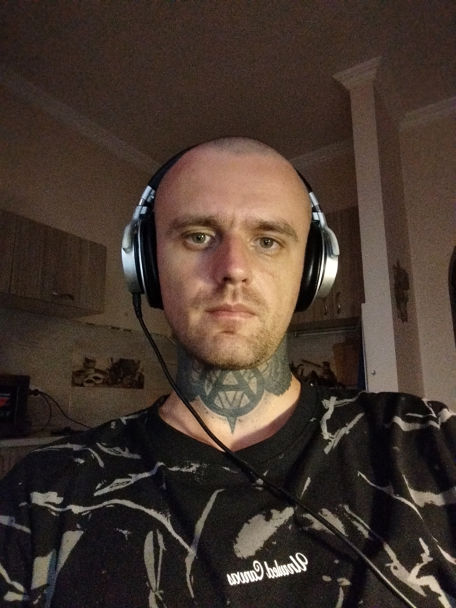 The image shows a close-up, waist-up shot of a bald, white male wearing over-ear headphones and a black t-shirt with a white, abstract pattern. He has a tattoo on his neck. The man is looking directly at the camera. The background appears to be a room with cabinets and a column, suggesting an indoor environment. The lighting is dim, and the image quality is grainy. The location is identified as Batumi, Georgia.