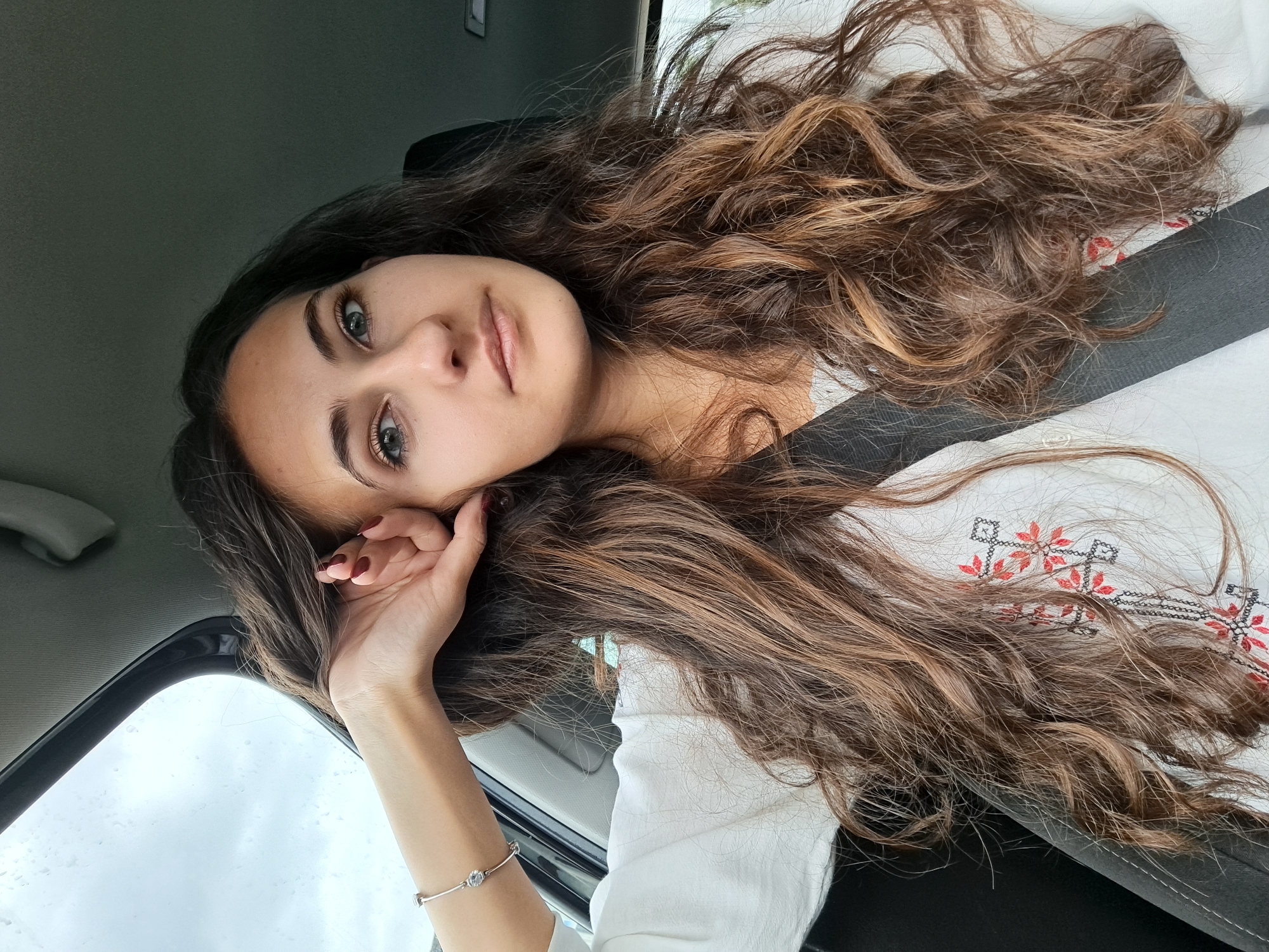 A young woman with long, wavy brown hair with blonde highlights is positioned in the front seat of a car. She is wearing a white blouse with embroidered red and black floral patterns. She has a black seatbelt over her right shoulder. Her left hand is resting on her face, and she is wearing a bracelet on her left wrist. The background shows the car's interior, including the dashboard, window, and headliner. Through the window, the sky is visible. The location is likely in Sofia, Bulgaria.