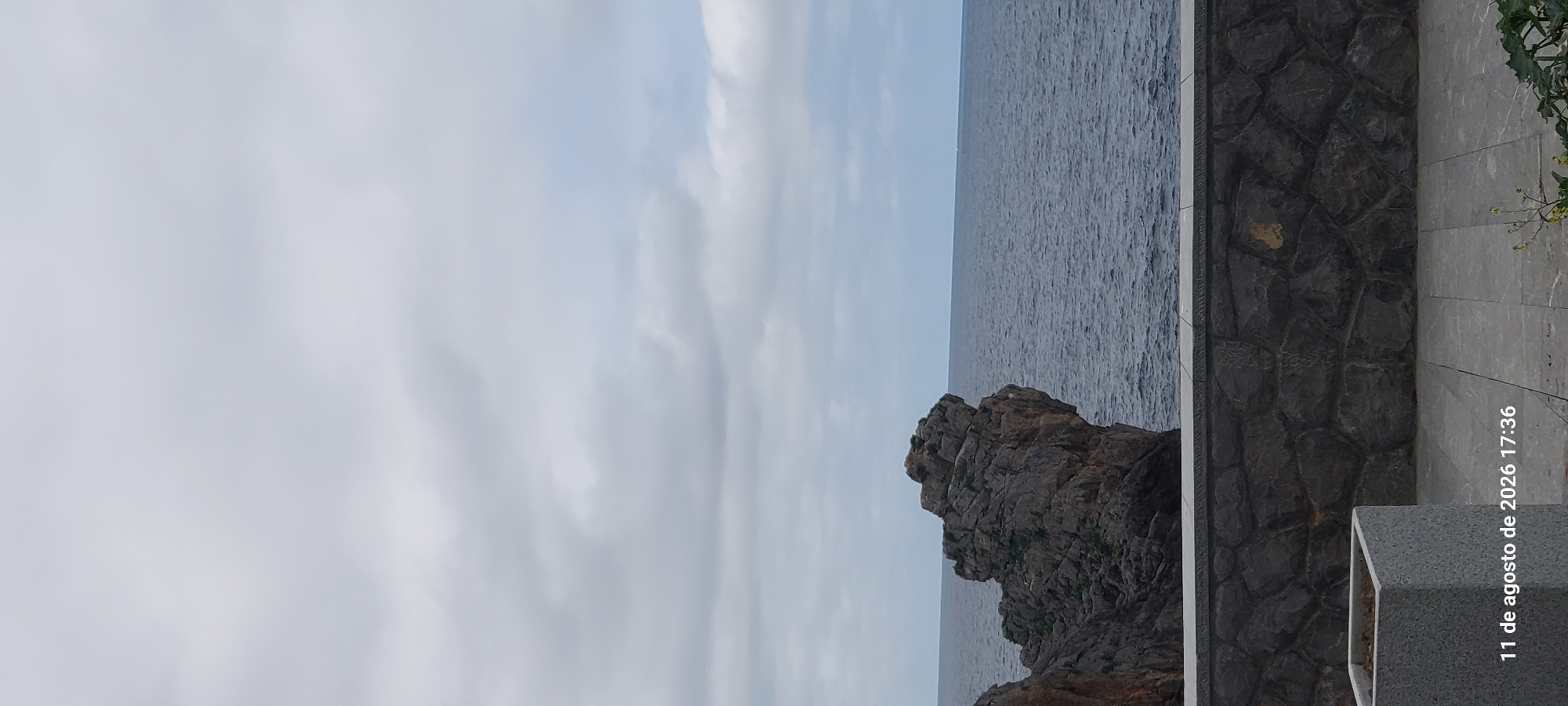 The image depicts a cloudy day over a rocky coastline and the sea. In the foreground, there is a low stone wall with a decorative border, and a square planter box to the left. The ground is paved with large rectangular tiles. The sea is a deep blue, with small waves. A large, rugged rock formation juts out into the sea on the left side of the image. The sky is overcast with gray clouds. The date and time stamp "11 de agosto de 2026 17:36" is visible in the bottom left corner. This scene is consistent with the coastal landscape of Castro-Urdiales, Spain.