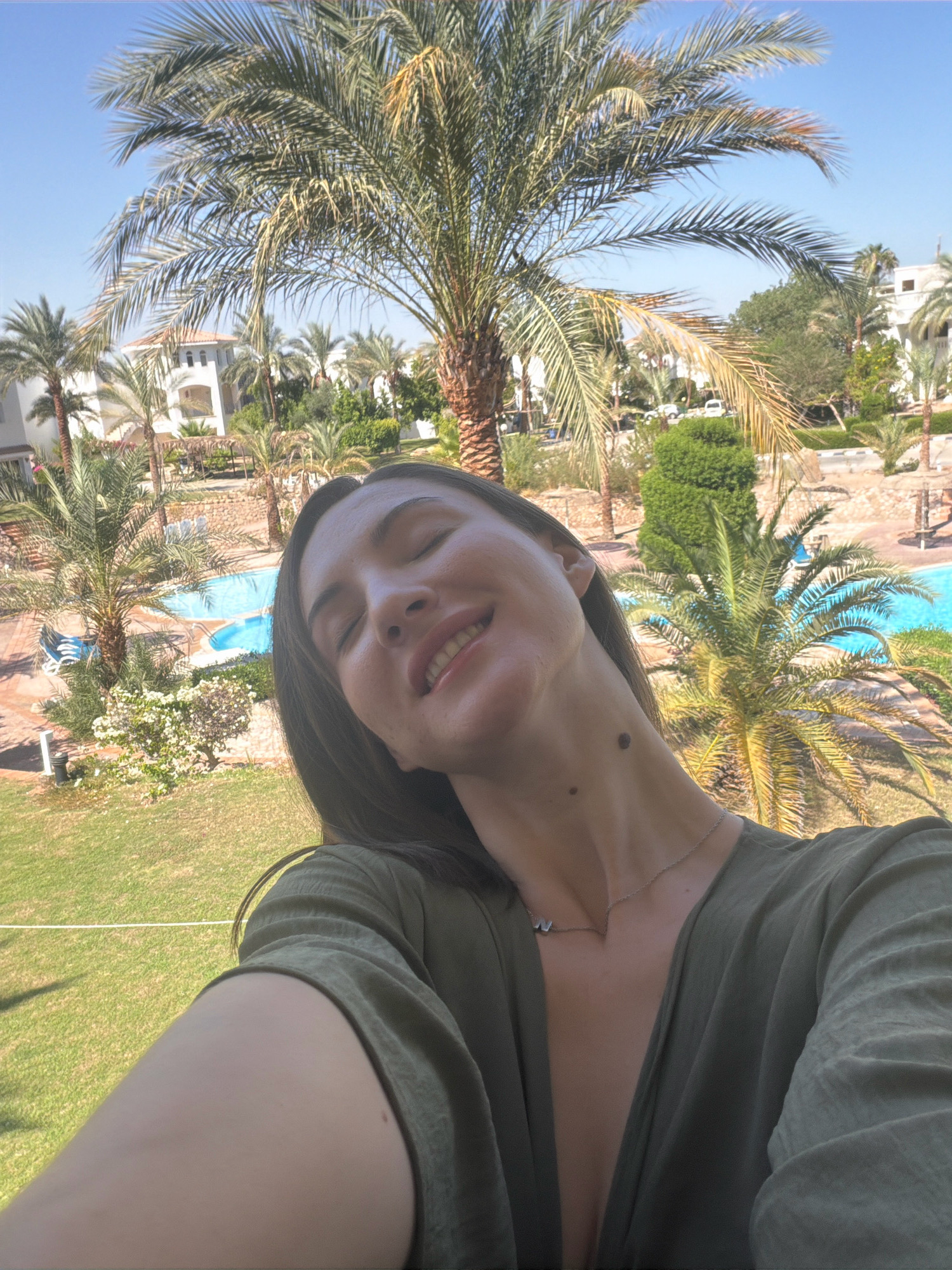 A woman with closed eyes and a wide smile is taking a selfie with a body of water and palm trees in the background. She is wearing a green dress and a silver necklace with a "W" pendant. The location is Sharm El Sheikh, Egypt. The woman is outdoors, in what appears to be a resort or hotel area. The sky is clear and blue, and the sun is shining. The scene is bright and vibrant, with lush green vegetation and the turquoise water of a swimming pool. The woman's hair is dark and falls around her shoulders. She has a mole on her neck.