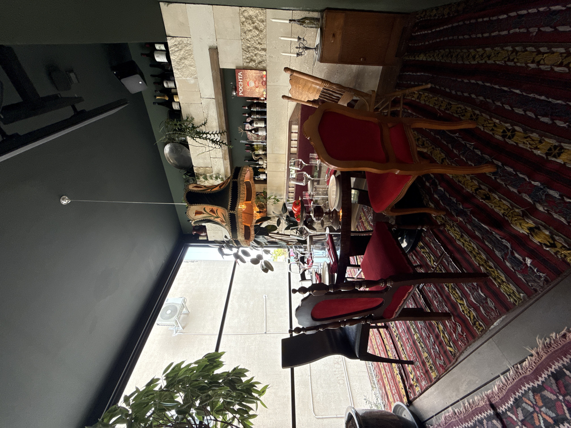 The image depicts the interior of a restaurant or dining area, possibly located in Limassol, Cyprus. The scene is set around a circular dining table laid with white plates, red napkins, wine glasses, and silverware. A centerpiece with red fruits is visible on the table. Several chairs surround the table: two dark-colored chairs, two chairs with red upholstered seats and wooden frames, and a wooden chair. In the background, shelves are lined with wine bottles and decorative items, including a sign that reads "POCHTA". A distinctive lampshade with a floral pattern hangs over the table. Large windows offer a view of an outdoor area with building exteriors and greenery. The flooring is covered with vibrantly patterned rugs. The overall atmosphere suggests a well-decorated and possibly upscale dining establishment.