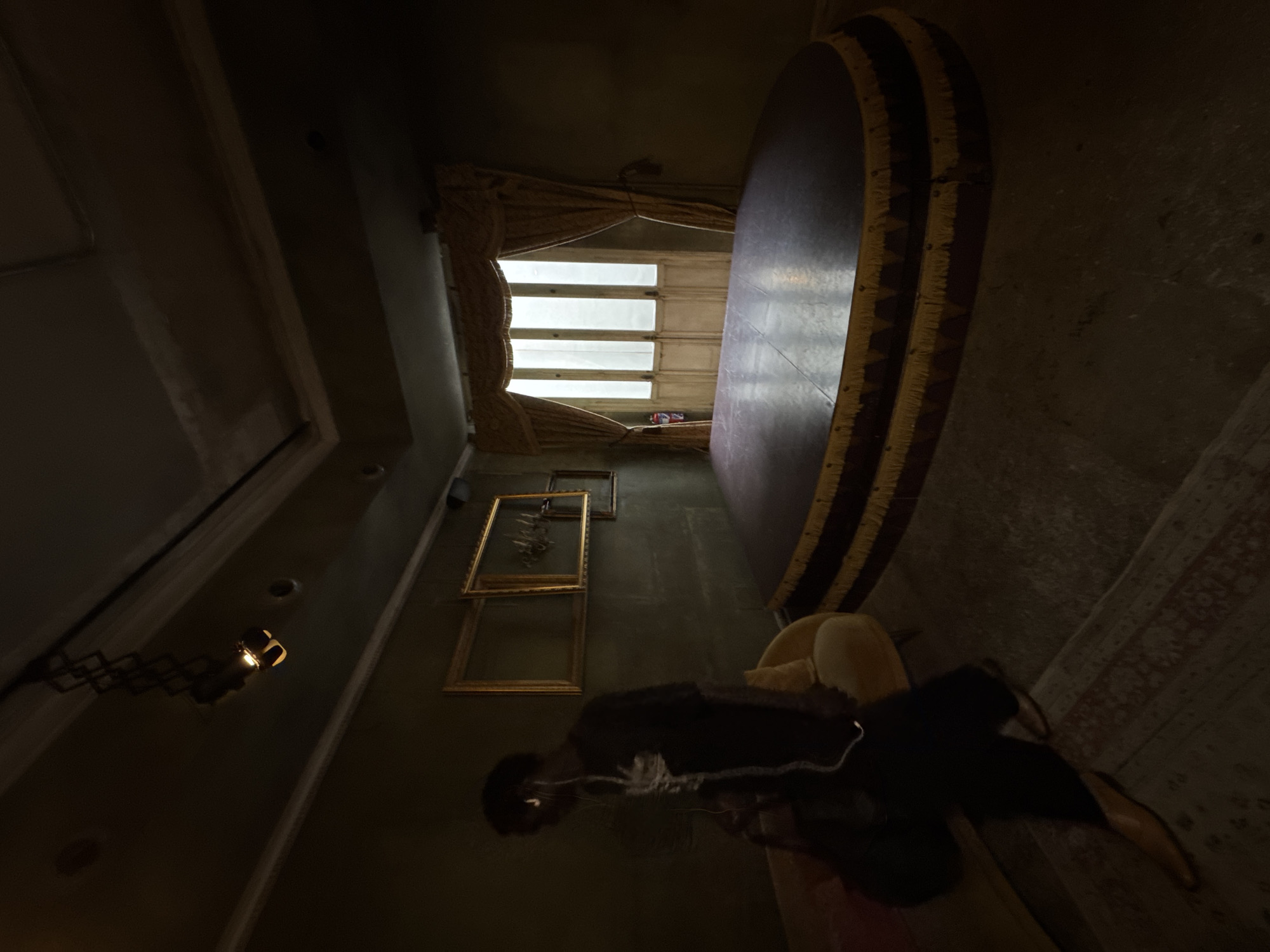 The image shows a dimly lit, ornately decorated room, captured from a low angle and rotated 90 degrees clockwise from its natural orientation. In the foreground, a person is visible, wearing a dark top and white earbuds, standing or sitting on a reflective dark floor.
On the left side of the room (if rotated back to a standard view), a large window with horizontal panes is present, letting in bright white daylight. This window is framed by heavy, textured curtains in a tan or gold color. Opposite the window, on the right wall, is a prominent, large, dark circular object with a distinctive patterned edge featuring yellow and dark brown fringes, possibly a decorative wall panel or a large round table viewed from an unusual angle.
Behind the person, on the same wall as the window, several empty gold-colored picture frames are hung, some overlapping. A wall-mounted light fixture with a geometric arm casts a warm glow on the far left wall. The floor exhibits both a dark, reflective surface and a patterned rug in reddish and beige tones. The walls appear to be a dark, muted color like dark green or grey.
The bright light from the window contrasts sharply with the overall dimness of the interior, suggesting either late daylight hours or a room designed to be dark. The scene depicts an indoor setting, potentially in Madrid, Spain, based on the provided context. No specific activity beyond the person's presence is evident, and no text is visible.