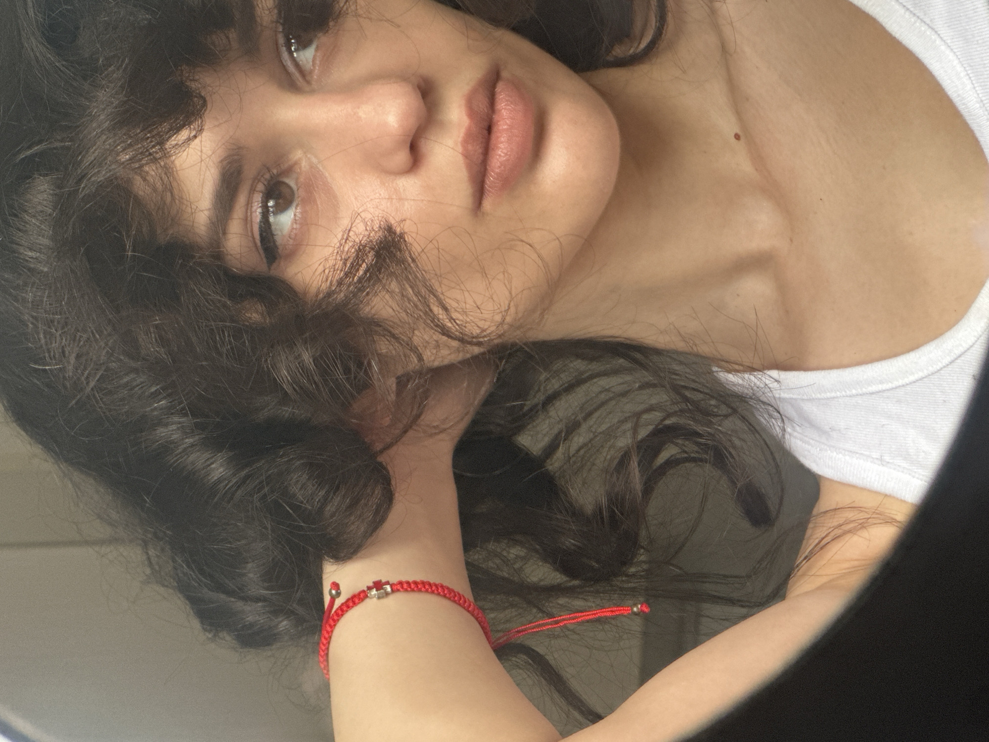 The image is a close-up, waist-up shot of a young woman with dark, curly hair, taken indoors. She is wearing a white tank top and a red string bracelet with a small, silver charm on her left wrist. Her left arm is raised, with her hand resting on the back of her head. She is looking to her right, with her head tilted slightly. The background is softly blurred and appears to be a neutral-colored wall. There is no indication of the city of Sofia, Bulgaria, or any other specific geographical location. The lighting is natural, coming from the right side of the frame, casting soft shadows on her face and neck. The image is composed to highlight the woman's features and her casual, relaxed posture.