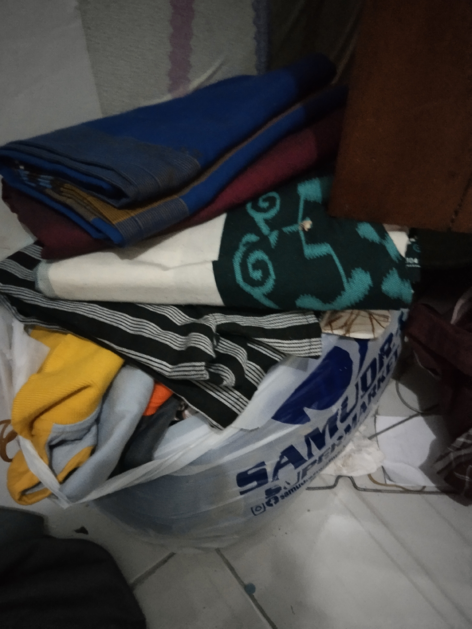 The image depicts a pile of folded clothing inside a clear plastic bag, which is placed on a tiled floor. The clothing items include what appear to be blankets or large pieces of fabric in various colors such as blue, maroon, white with green patterns, black and white stripes, and yellow. The plastic bag has "SAMURAI MARKET" printed on it in large blue letters. The scene is indoors, with a wall visible in the background. The lighting is dim. The floor tiles are light-colored and arranged in a grid pattern. The overall impression is that of laundry or stored textiles. The location is identified as Buntalan, Indonesia.