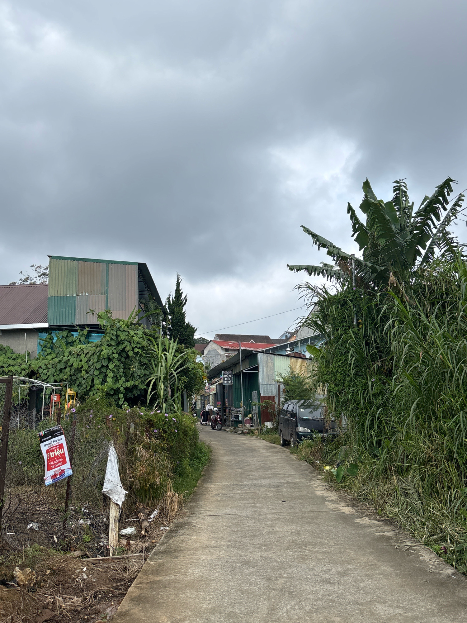 A vertical, slightly low-angle shot captures a narrow, concrete road leading into a residential area under a cloudy, overcast sky. The road is flanked by overgrown vegetation on the right and a mix of buildings and foliage on the left.
On the left side of the road, the foreground shows a dirt embankment with some debris, including a white plastic bag tied to a wooden post. A red and white sign, possibly an advertisement, is attached to a metal fence. Behind the fence, there is lush green foliage and several plants, including what appears to be a small, multi-story building with a corrugated metal roof and walls of various colors. Further back, other buildings with pitched roofs are visible, along with a tall, conical evergreen tree.
The right side of the road is dominated by dense, tall green grass and a large banana tree with its distinctive large leaves. A black minivan is parked partially obscured by the vegetation on the right side of the road.
In the mid-ground, the road continues to curve slightly. A few motorcycles are parked in front of buildings on the left, and a few individuals are visible in the distance. The buildings on this side appear to be smaller, some with corrugated metal siding and others with more traditional roofing materials. One building has a sign with Vietnamese text that includes "BÚN BÒ HUẾ."
The sky is a uniform grey, suggesting a cloudy or possibly rainy day. The overall impression is of a modest, perhaps developing, urban or semi-urban environment.