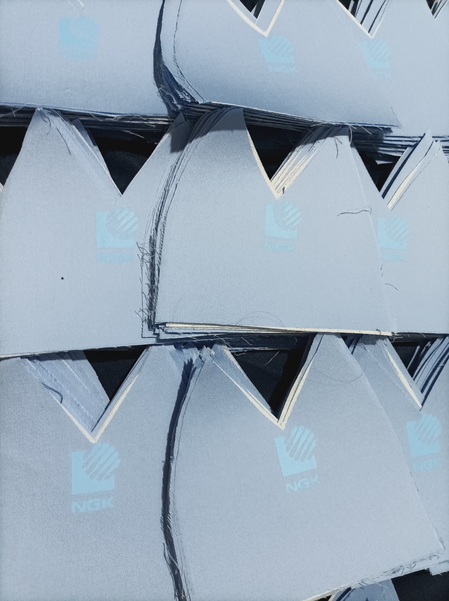 The image displays a stack of light blue, triangular fabric pieces, possibly cutouts for manufacturing. Each piece bears a subtle light blue logo that appears to be "NGK" with a stylized graphic above the text. The fabric has a visible texture of fine horizontal lines. The pieces are arranged in rows, overlapping and creating a layered effect. Some of the edges show fraying or stitching. The background is dark, providing a contrast to the fabric. The lighting is even, and there are no discernible shadows that would indicate a specific time of day or light source. The image appears to be a close-up, studio shot of the materials. This image does not contain any recognizable landmarks or elements that would allow for the identification of a specific location in West Jakarta, Indonesia.