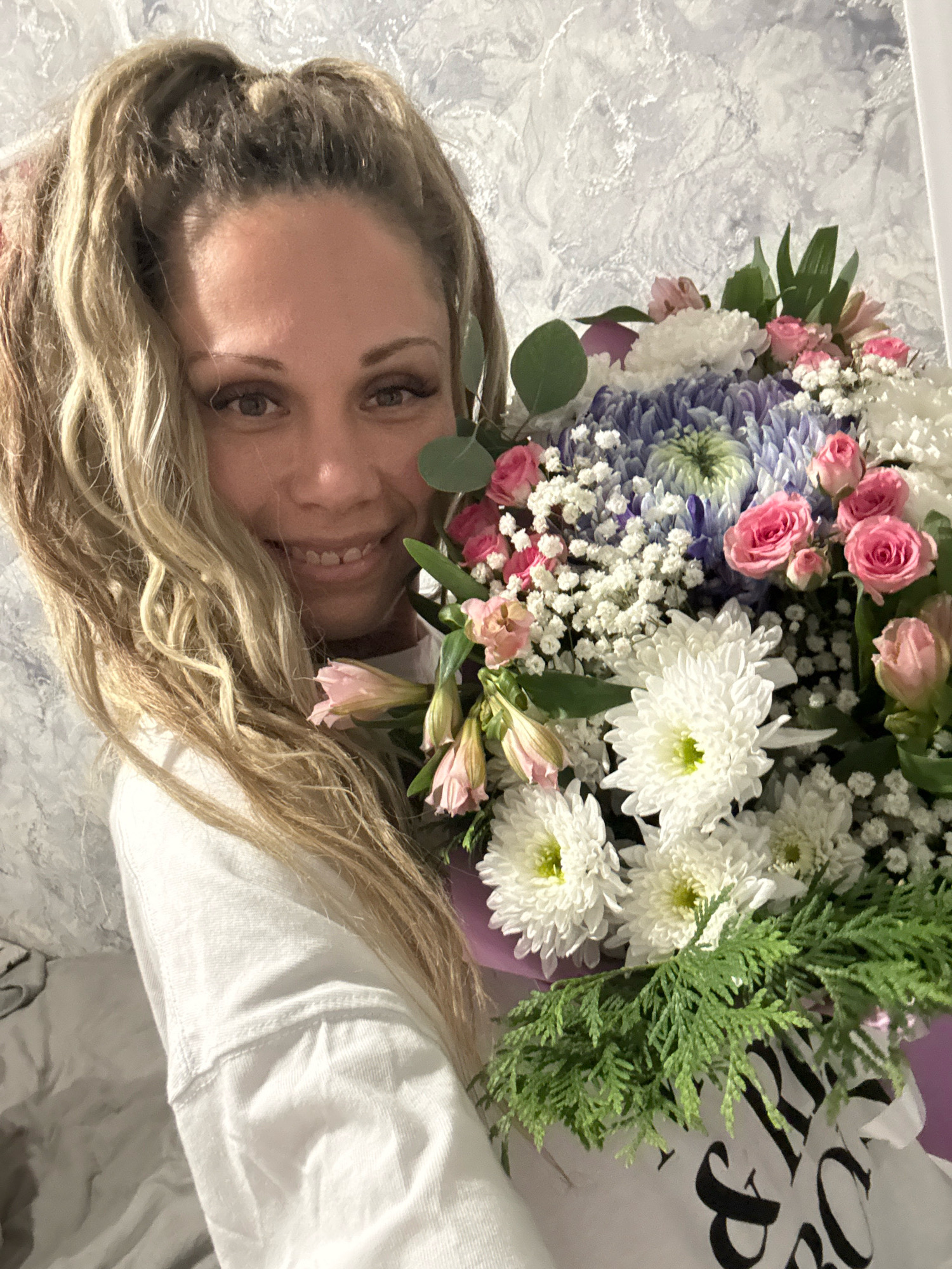 A Caucasian woman with blonde, curly, high ponytail hair smiles while holding a large bouquet of flowers. She is wearing a white sweatshirt. The bouquet consists of white and pink roses, blue and white chrysanthemums, and baby's breath. The background is a textured light-colored wall. The scene appears to be indoors. The city of Bor, Russia is not depicted in this image.