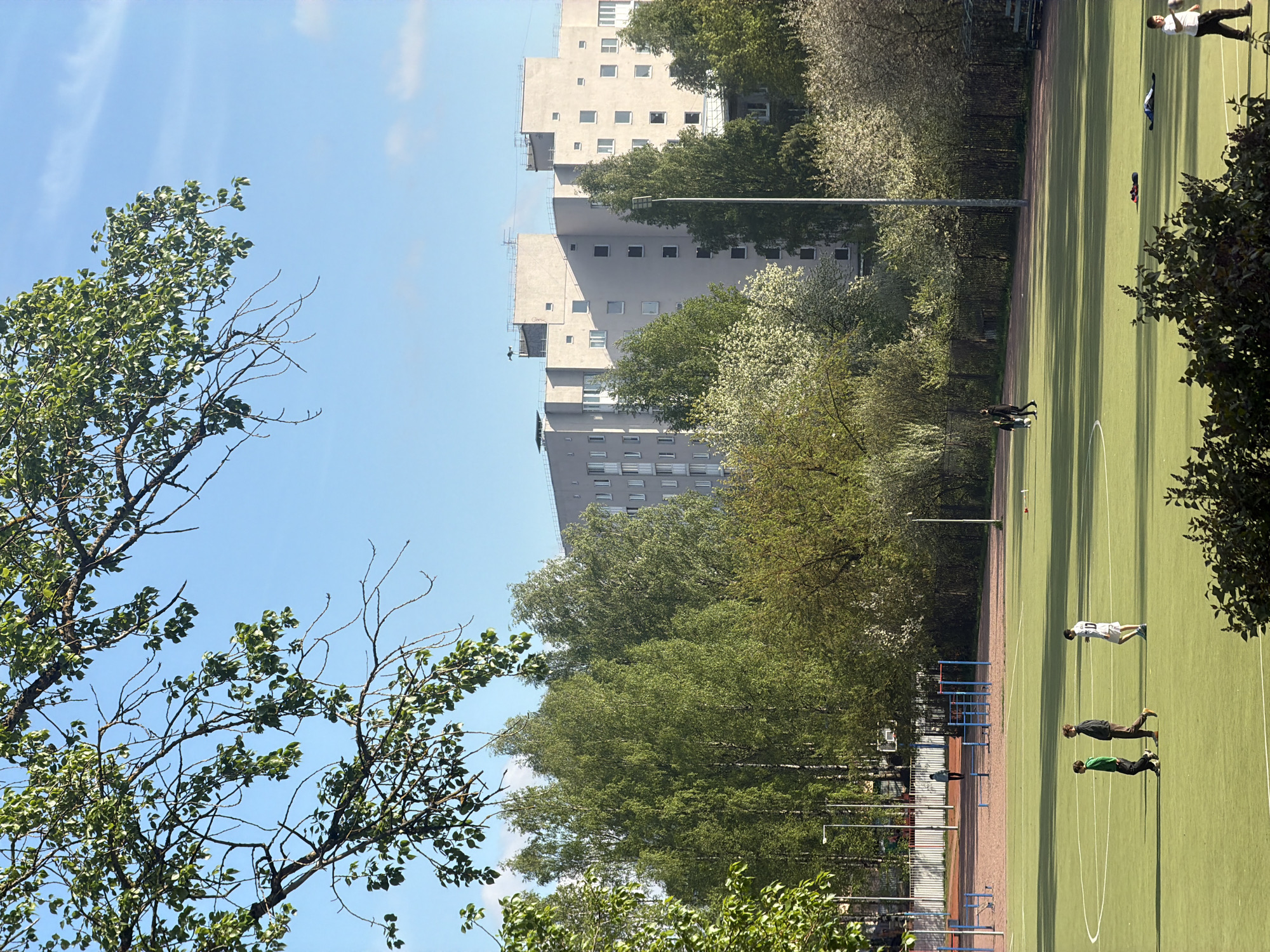 The image presents a view of Tokkari-Lend, Russia, featuring a sports field bordered by a line of trees and a tall apartment building in the background. The sky is blue with some clouds. In the lower left corner, a tree with green leaves and bare branches frames the shot.
On the green field, several people are present. Towards the right side, there is an individual lying face down on the ground, and nearby is another person who seems to be standing or walking. Further to the left, a line of two people appears to be jogging. There are also some blue training hurdles near the edge of the field, adjacent to a fence.
The backdrop showcases the apartment building, its gray exterior punctuated by windows. Trees partially obscure the lower floors of the building. A street light pole is also visible among the trees. The overall scene suggests a combination of urban and natural elements in the location.