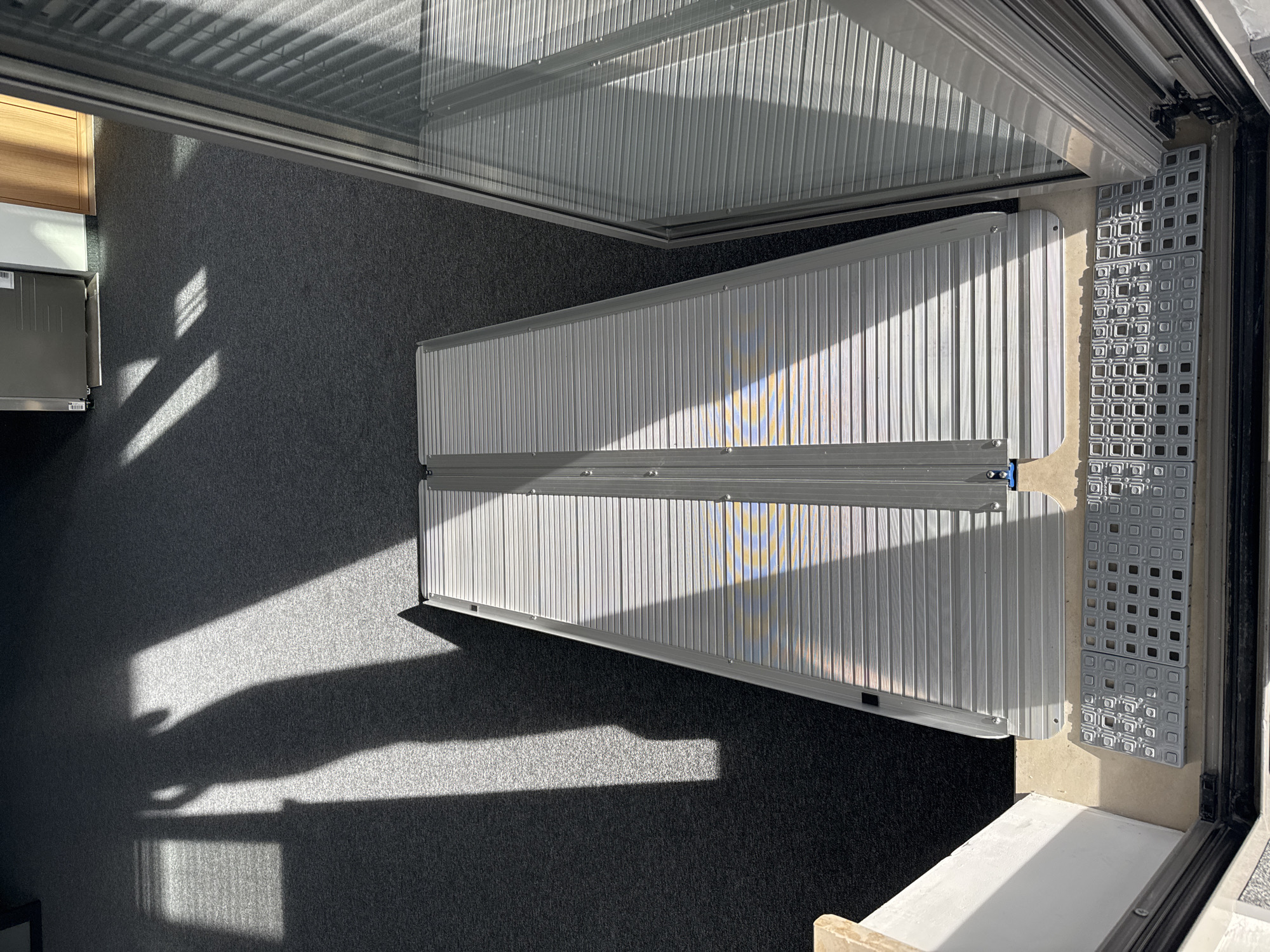 The image shows an interior space with a gray carpeted floor. A collapsible aluminum ramp extends from the doorway into the room. Sunlight streams in through the doorway, casting shadows on the floor and ramp. The doorway features a gray, square-patterned landing pad, and a track for a sliding door. Some wooden furniture is partially visible in the background. There's no clear indication of the location being Sofia, Bulgaria.