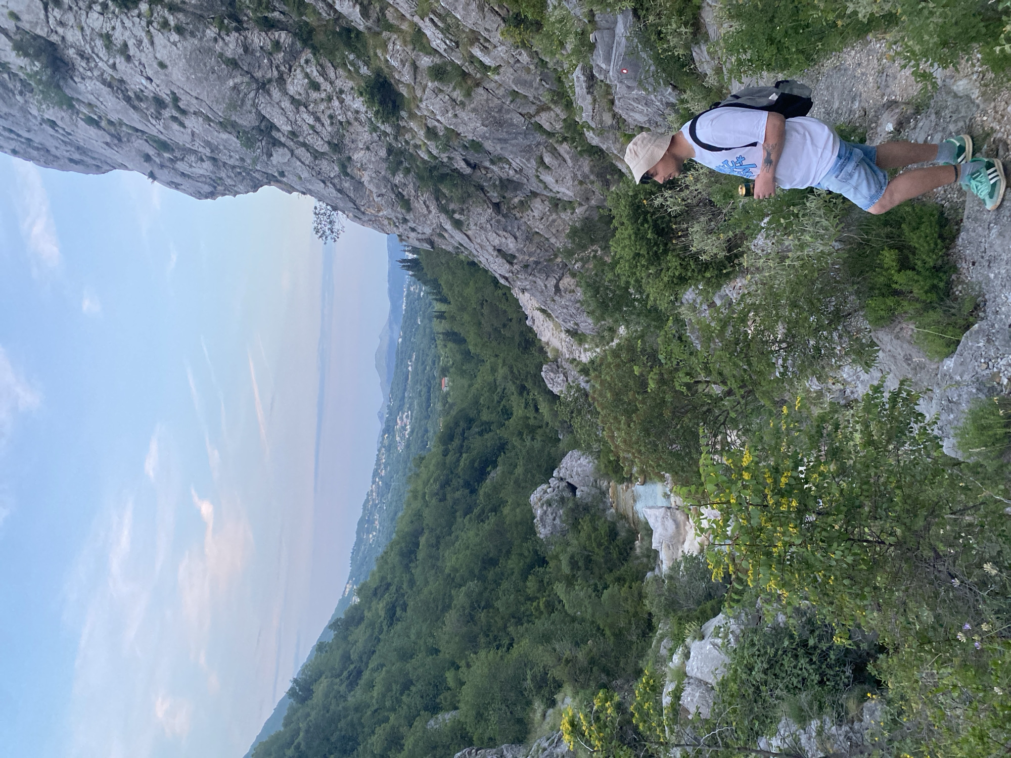 The image, set in Zupci, Montenegro, depicts a male hiker on a narrow, rocky mountain path during late afternoon or early evening. The hiker wears a white t-shirt, light blue shorts, a beige bucket hat, and light green sneakers, carrying a dark backpack. He is looking down at a phone he holds in his hand.
Beside him, a towering, rugged grey cliff face rises sharply. Below him, the steep slope is densely covered with vibrant green trees and bushes. A distinctive light-colored rock formation, possibly a mineral deposit or part of a spring, is visible further down the slope amidst the trees. In the distance, a tree-filled valley with scattered houses extends to a vast body of water, likely the Adriatic Sea, beneath a soft blue sky with faint clouds. A small red circular trail marker is faintly visible on a rock face above the path. The overall impression is of an adventurous trek through a dramatic, lush mountain landscape. No readable text is visible.