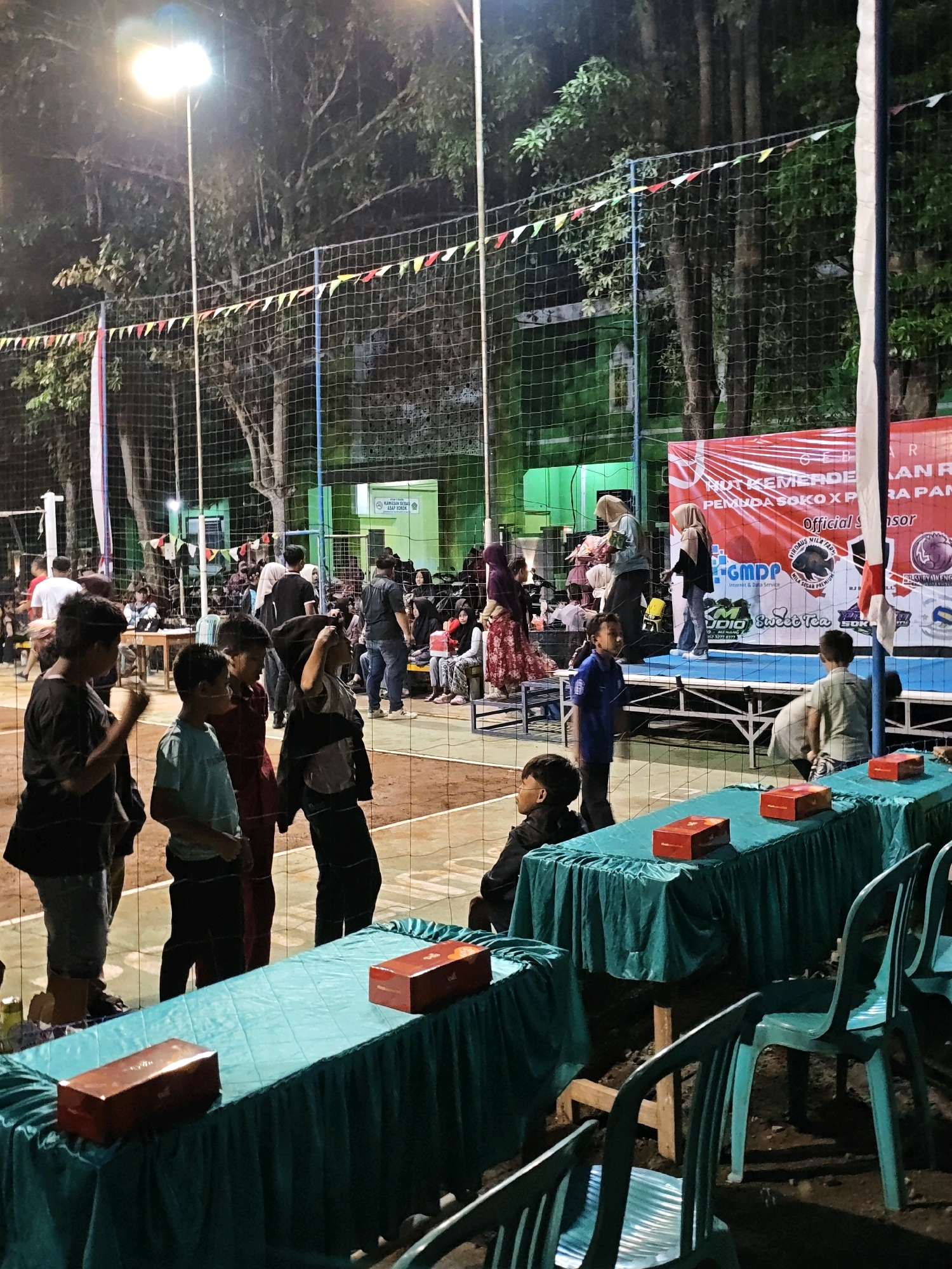 The image depicts an outdoor event at night, likely a sports tournament, on a volleyball court in Pagu, Indonesia. In the foreground, two tables covered with teal tablecloths are set up, each holding two red gift boxes. Several turquoise plastic chairs are arranged in front of the tables. A volleyball net is prominently visible, stretching across the court. Behind the net, a group of people, including children and adults, are gathered. Some are standing, while others are seated on bleachers or on the ground. A stage is set up with a large red banner that has text and logos. The banner appears to be for an event celebrating Indonesian Independence Day ("HUT KEMERDEKAN"). A street light illuminates the scene from the top left, casting a bright glow. Trees with dense foliage surround the court, and some string lights are visible. The overall atmosphere suggests a community gathering and celebration.