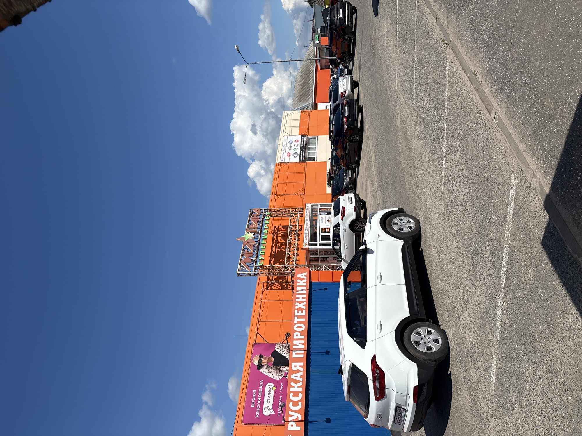 In Sharya, Russia, a commercial building with an orange facade displays signage for "РУССКАЯ ПИРОТЕХНИКА" (Russian Fireworks) and a supermarket. A billboard features a woman promoting women's outerwear. A parking lot in front of the building is filled with parked vehicles, including white SUVs, black sedans, and other cars. The sky is blue with scattered white clouds. The scene appears to be a sunny day in a commercial district.