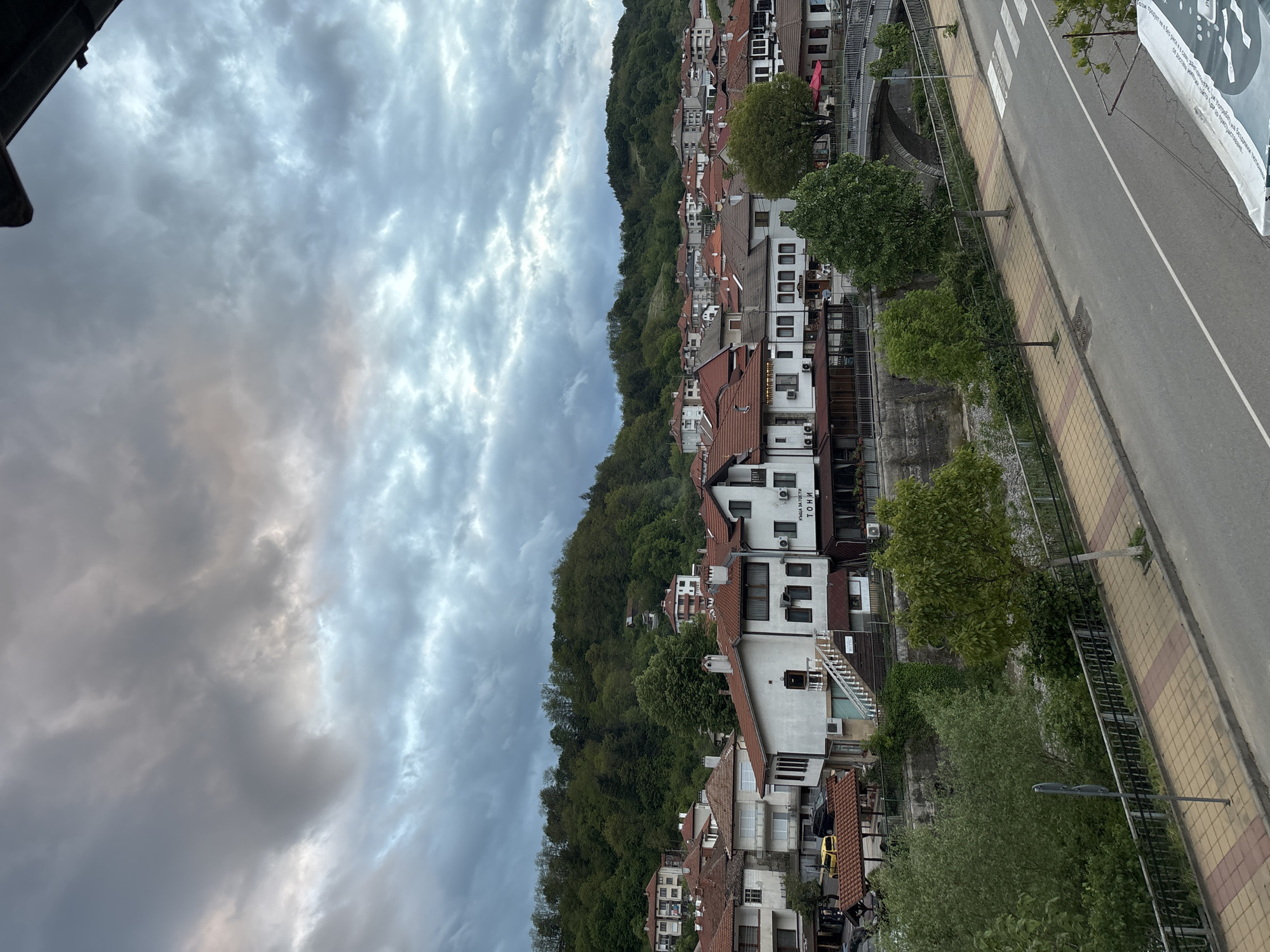 The image depicts a hillside town under a cloudy sky. The town, likely Zlatograd, Bulgaria, is built along a hillside covered in trees. Buildings are mainly white with red-tiled roofs. A road runs parallel to the buildings with trees lining the sidewalk. The sidewalk is made of yellow and red pavers. A billboard is partially visible on the right side of the image.