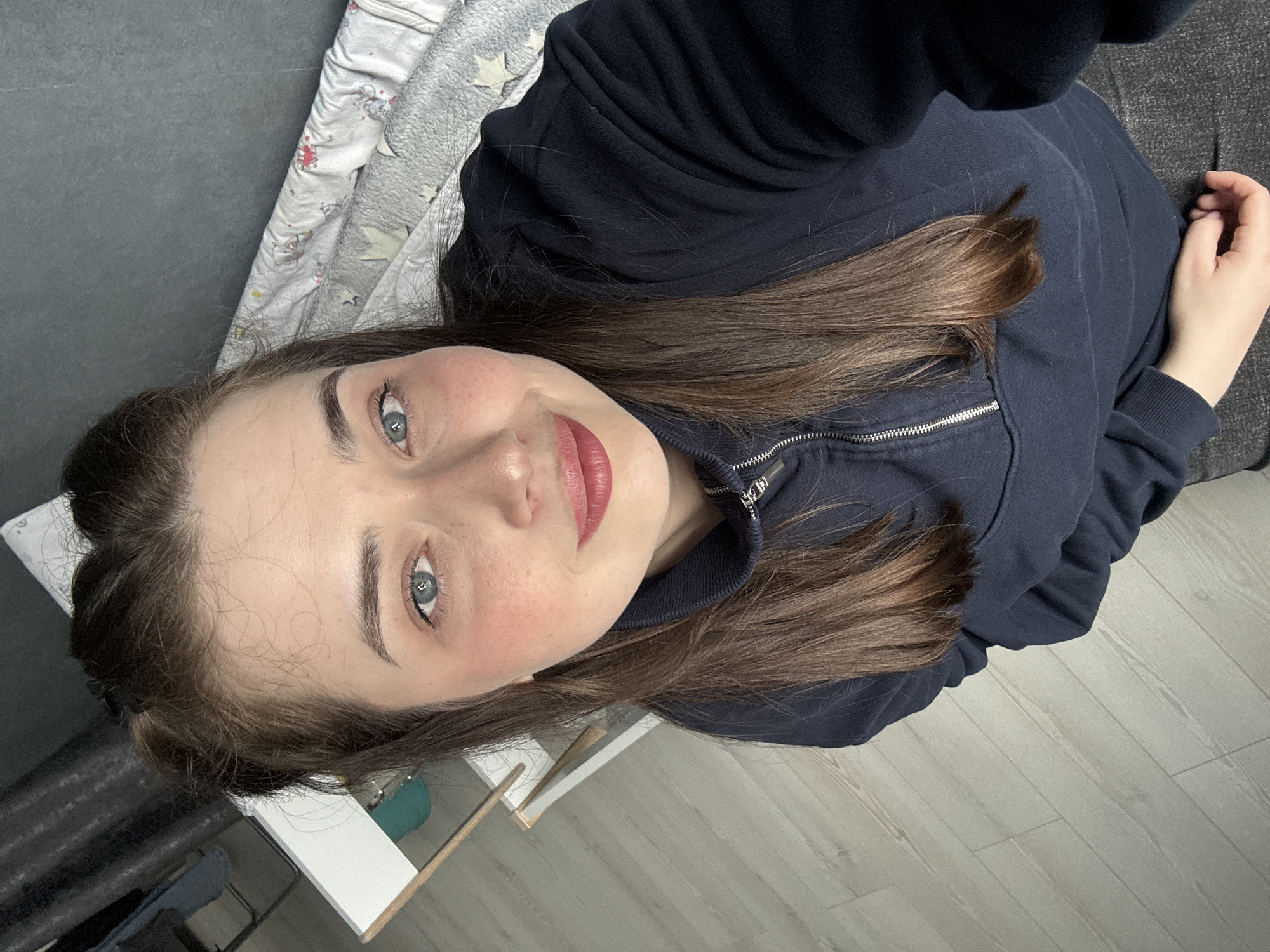 A young woman with fair skin, light blue eyes, and brown hair is lying down and looking up at the camera. She is wearing a navy blue zip-up jacket. Her hair is partially pulled back, and she has lipstick on. Her right hand is visible near the top right of the frame. The background includes a bed with a patterned blanket and a gray couch. A white desk with a green object underneath is visible on the left side of the frame. The floor appears to be light-colored laminate flooring. The location is likely indoors. It's unlikely any information can be gathered about it being in Chaikovsky, Russia from only this image.