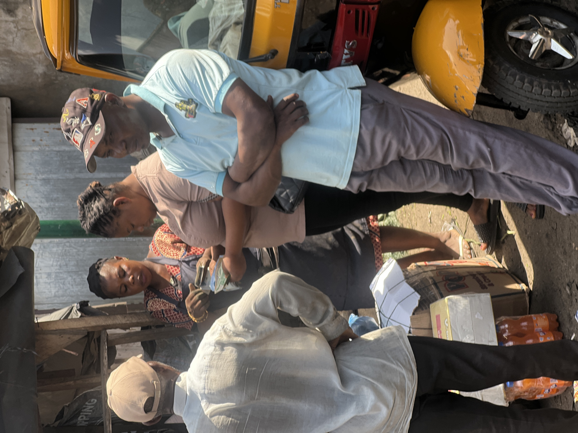 This image, set in Nkpor, Nigeria, captures a vibrant outdoor marketplace scene during bright daylight hours, indicated by strong shadows. Several individuals are engaged in what appears to be commercial activity.
In the foreground, a man with his back to the viewer wears a light-colored long-sleeved shirt and dark trousers, topped with a beige baseball cap. He is leaning forward, seemingly interacting with goods or another person at a stall. Next to him, a woman in a patterned top (red, brown, white) and dark skirt, adorned with gold bangles, is visible. She holds what appears to be money or small items, suggesting a transaction is taking place. Behind her, another woman in a light brown top and dark skirt observes the scene.
A prominent figure in the middle ground is a man in a light blue polo shirt and grey trousers, wearing a dark baseball cap embellished with various patches, including symbols resembling the Louis Vuitton logo. He stands with his arms crossed, his gaze directed downwards.
The setting is bustling, with a yellow Keke Napep (auto-rickshaw) partially visible on the left and right, featuring its distinctive yellow body and tire. A red container or crate marked with "SA..." is near the Keke. Various goods are stacked around the people, including cardboard boxes—one showing "VG"—and bundles of plastic bottles containing an orange-colored liquid, likely soda or juice. A wooden structure, possibly part of a market stall, is in the background. The ground appears to be a mix of dirt and pavement, characteristic of an open-air market. The overall impression is one of everyday commerce and interaction in a lively urban environment.