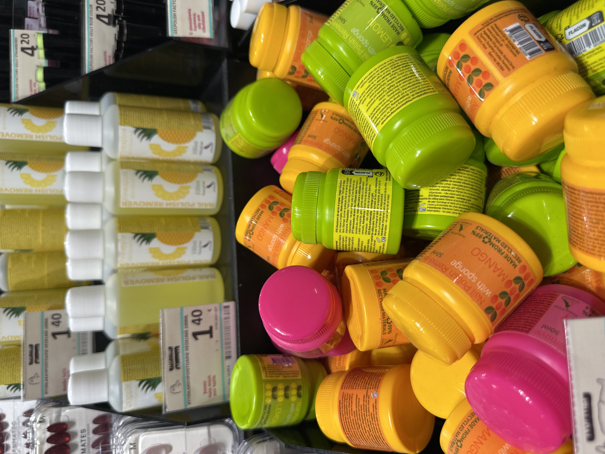 This is a close-up, eye-level shot of a retail display featuring multiple bottles and jars of nail polish remover. The products are predominantly in bright, saturated colors such as neon green, hot pink, and bright yellow. Many of the containers are small, round jars with prominent screw-on lids, while others are taller, slender bottles. The labels on the products indicate various scents like "Mango" and "Lemon," and some also state "Nail Polish Remover with Sponge." The background is a blurred array of more nail polish removers and other cosmetic products, with some pricing tags visible. The scene appears to be well-lit, suggesting an indoor retail environment.