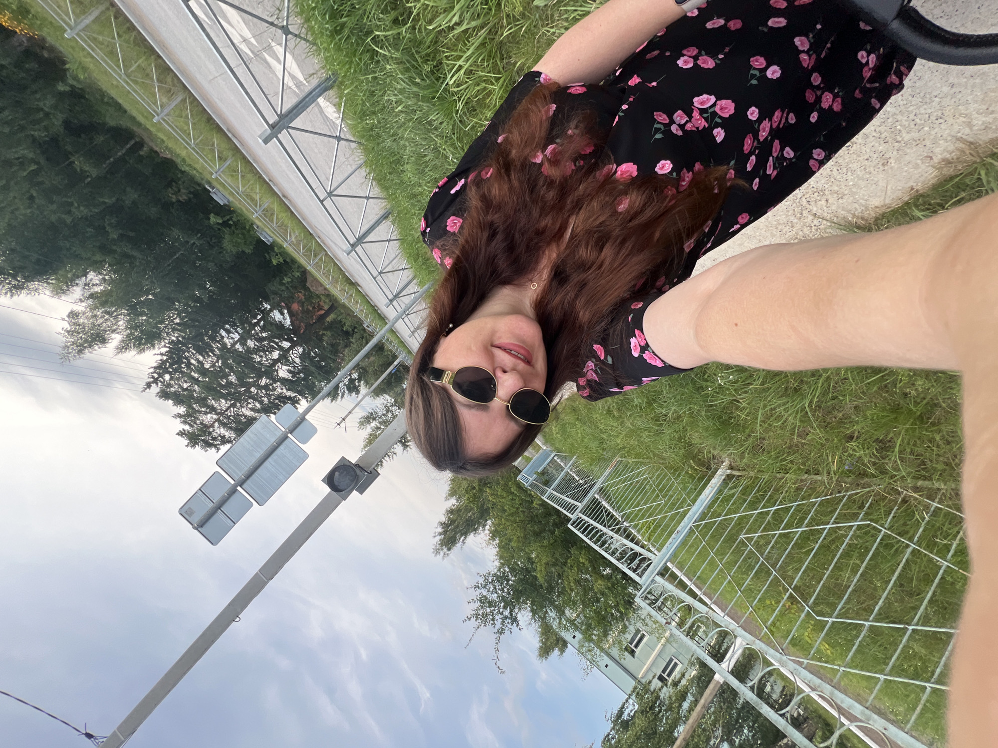 A young woman with long, dark, wavy hair and sunglasses is taking a selfie outdoors. She is wearing a black floral dress. She appears to be standing next to a metal fence and a grassy area, with a road and trees in the background. A traffic light and signs are visible on a pole. A building with windows can be seen further in the distance. The scene suggests an urban or suburban environment in Noviy, Russia, possibly near a road or park. The sky is partly cloudy.