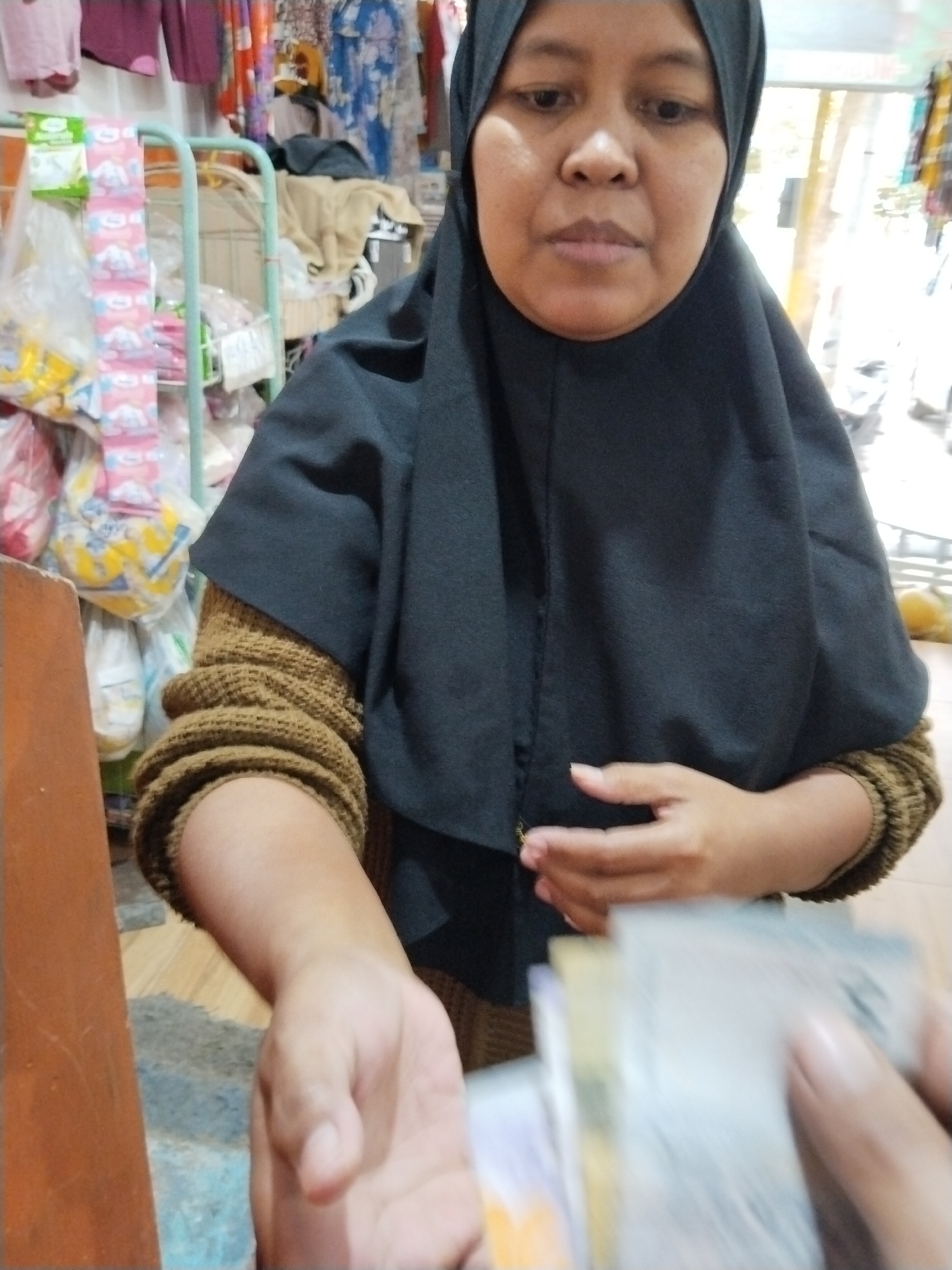 A medium shot captures a woman wearing a black hijab and a brown knitted sweater in what appears to be a small shop. Her right hand is extended, holding out a fan of banknotes. The background is cluttered with merchandise, including hanging clothes and packaged goods displayed on racks. The scene suggests a transaction is taking place, possibly a purchase or payment. The lighting is natural, indicating an indoor setting during daytime. The woman's facial expression is neutral, and her gaze is directed slightly downwards. The composition focuses on the interaction between the woman and the unseen person holding the camera, highlighting the exchange of money. The overall impression is one of everyday commerce.
