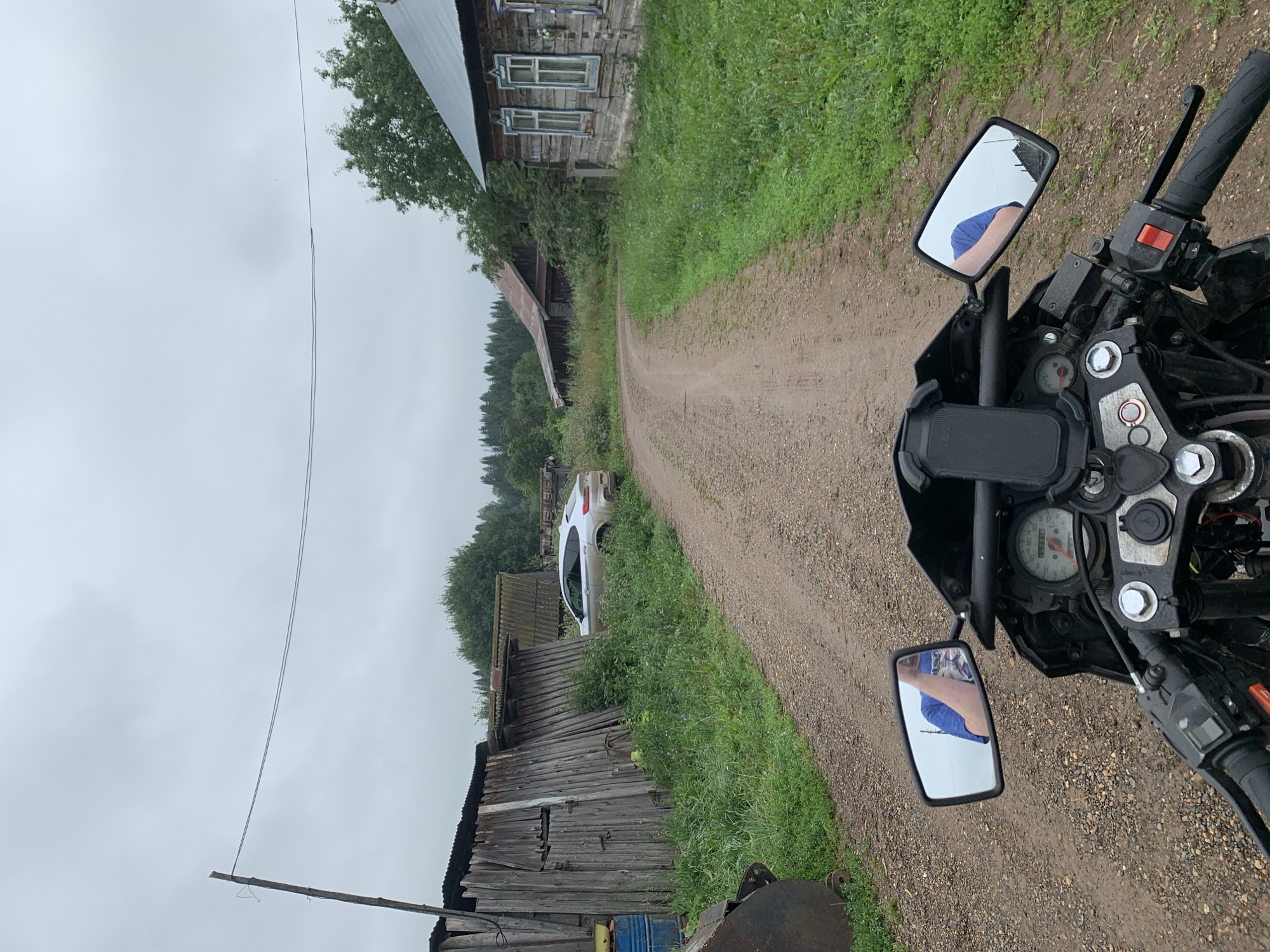 The image is taken from the perspective of a person riding a motorcycle. In the foreground, the handlebars and dashboard of the motorcycle are visible, including a speedometer and a phone mount. The rider's arms, clad in blue, are reflected in the motorcycle's mirrors. The motorcycle is on a dirt road that curves ahead. To the left of the road are dilapidated wooden structures and some overgrown greenery. Further down the road, a silver SUV is parked. On the right side of the road, a traditional wooden Russian house with blue window frames is situated. The background consists of trees and a cloudy sky. The overall scene depicts a rural or semi-rural setting in the village of Panteleevka, Russia, characterized by weathered buildings and natural elements.
