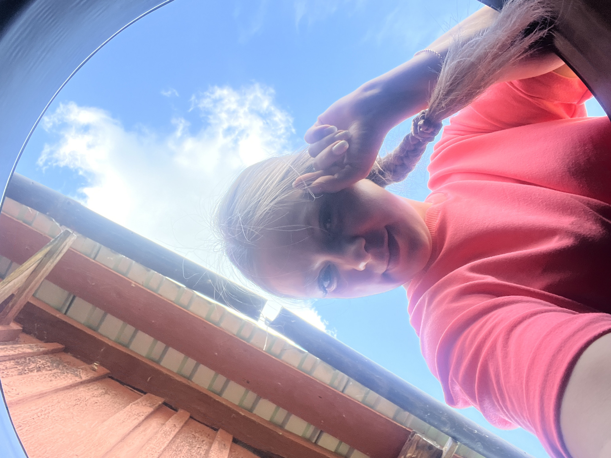 A low-angle, close-up shot captures a young woman with blonde hair in a braid, wearing a pink sweatshirt. She is looking up at the camera, with her right hand touching her head. Behind her, the sky is bright blue with scattered white clouds, and a section of a wooden roof structure is visible on the left. The image appears to be taken outdoors, possibly on a sunny day. The location within the city of Сухринское, Russia, is not discernible from the visual information provided.