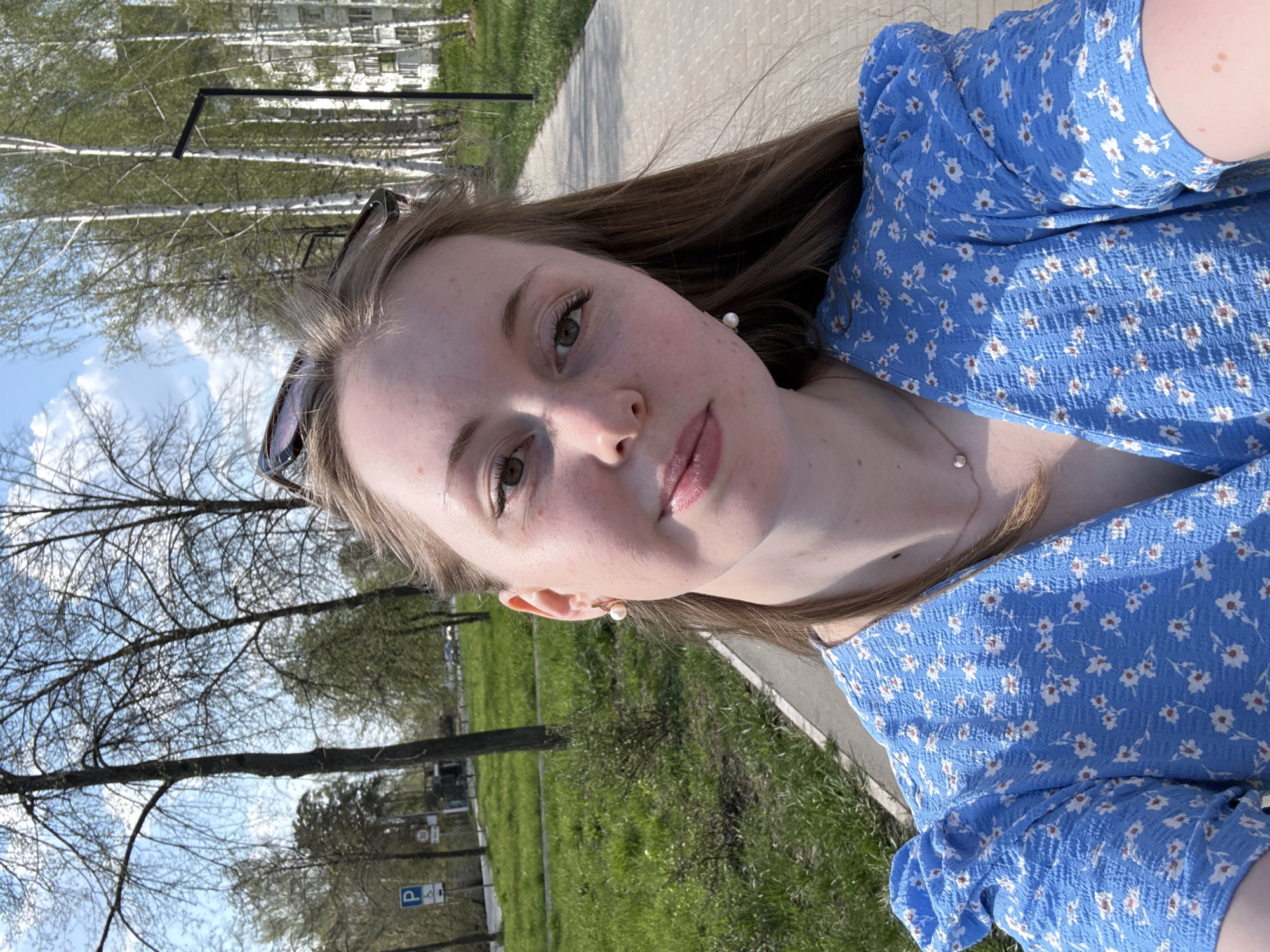 A selfie of a fair-skinned woman with auburn hair. She is wearing a blue dress with white floral patterns. She has a necklace and pearl earrings. Sunglasses are perched on her head. The background shows a park or green space with trees, grass, and a paved pathway. There is a building visible in the background. The location appears to be in a city, but the specific city and details cannot be determined from the image.