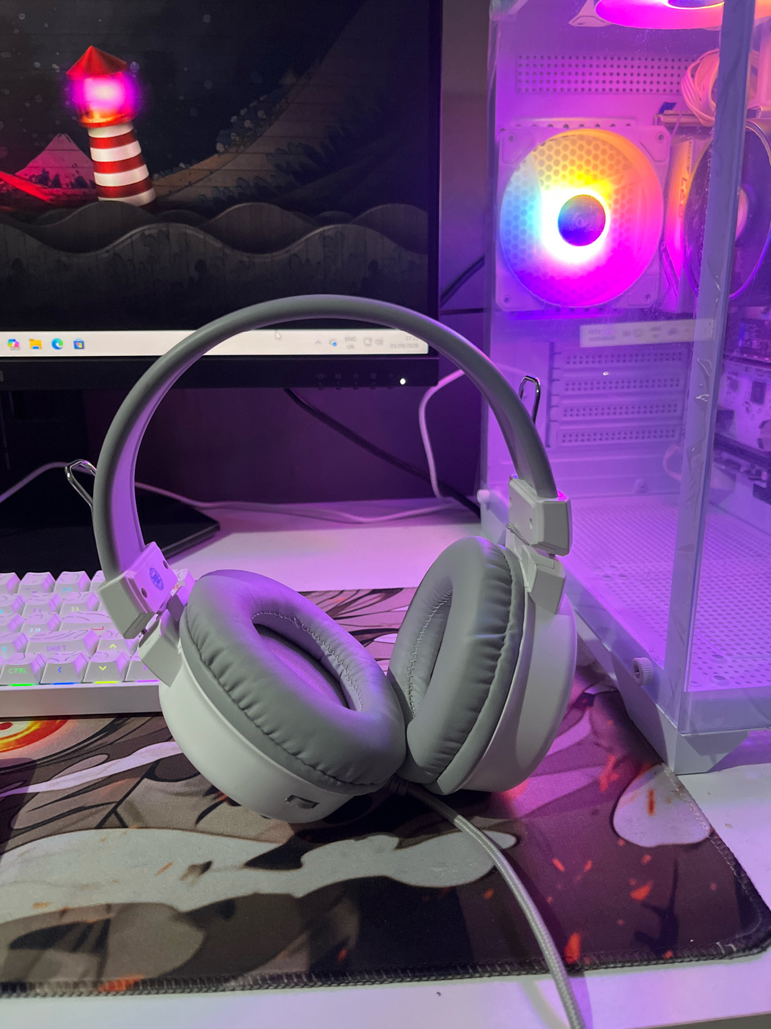 In the foreground of the image, a pair of white and gray headphones rests on a patterned desk mat. The headphones have a thick, gray headband and soft gray earcups. To the left of the headphones is a white mechanical keyboard with backlit keys displaying various colors, including green, blue, and yellow.
Behind the keyboard, a computer monitor displays a dark background with a stylized image of a lighthouse on a wavy sea. The lighthouse is red and white striped with a glowing pink top. The monitor itself has a thin bezel and appears to be a flat-screen display.
To the right of the headphones and keyboard, a portion of a computer tower is visible. It has a transparent side panel, revealing internal components. A large, circular fan within the computer is illuminated with multicolored LED lights, creating a vibrant, rainbow effect. The computer tower and surrounding area are bathed in a purple light, likely from the computer's internal lighting.
The overall setting appears to be a desk or workstation. The lighting is predominantly artificial, with the purple glow from the computer influencing the scene. The image does not provide any specific geographical markers to determine a location within the city of Cimahi, Indonesia.