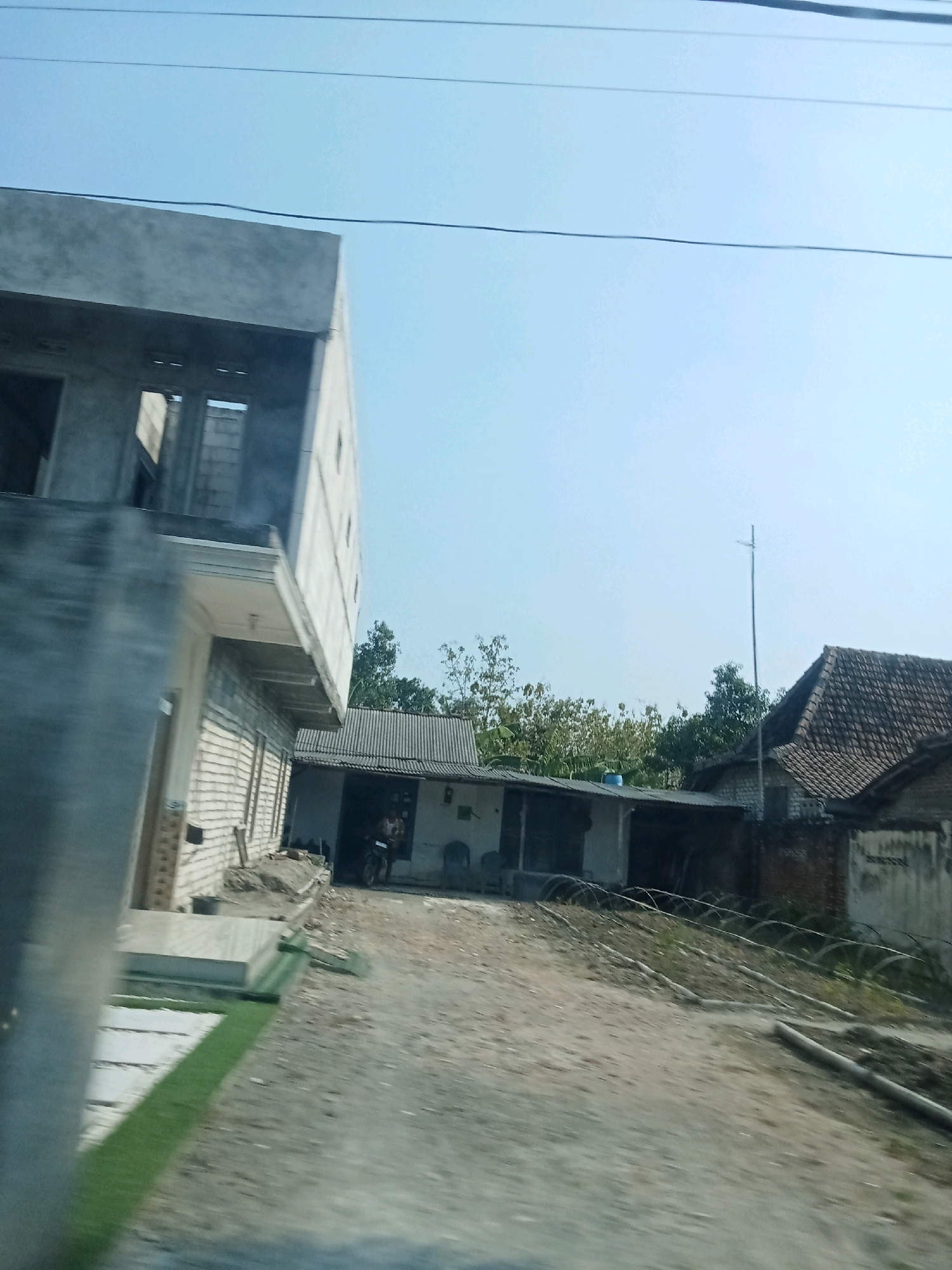 The image depicts a scene in Sumberringin, Indonesia, on a clear day. In the foreground, a partially constructed building with grey concrete walls and exposed brickwork is visible on the left. Adjacent to it, a lower structure with a tiled roof extends horizontally. A dirt path or road runs between these buildings and into the background, where more houses with tiled roofs and trees can be seen. There is a single person on a motorcycle in the open doorway of the lower structure. The overall impression is one of ongoing construction and residential development in a semi-urban or rural setting.
