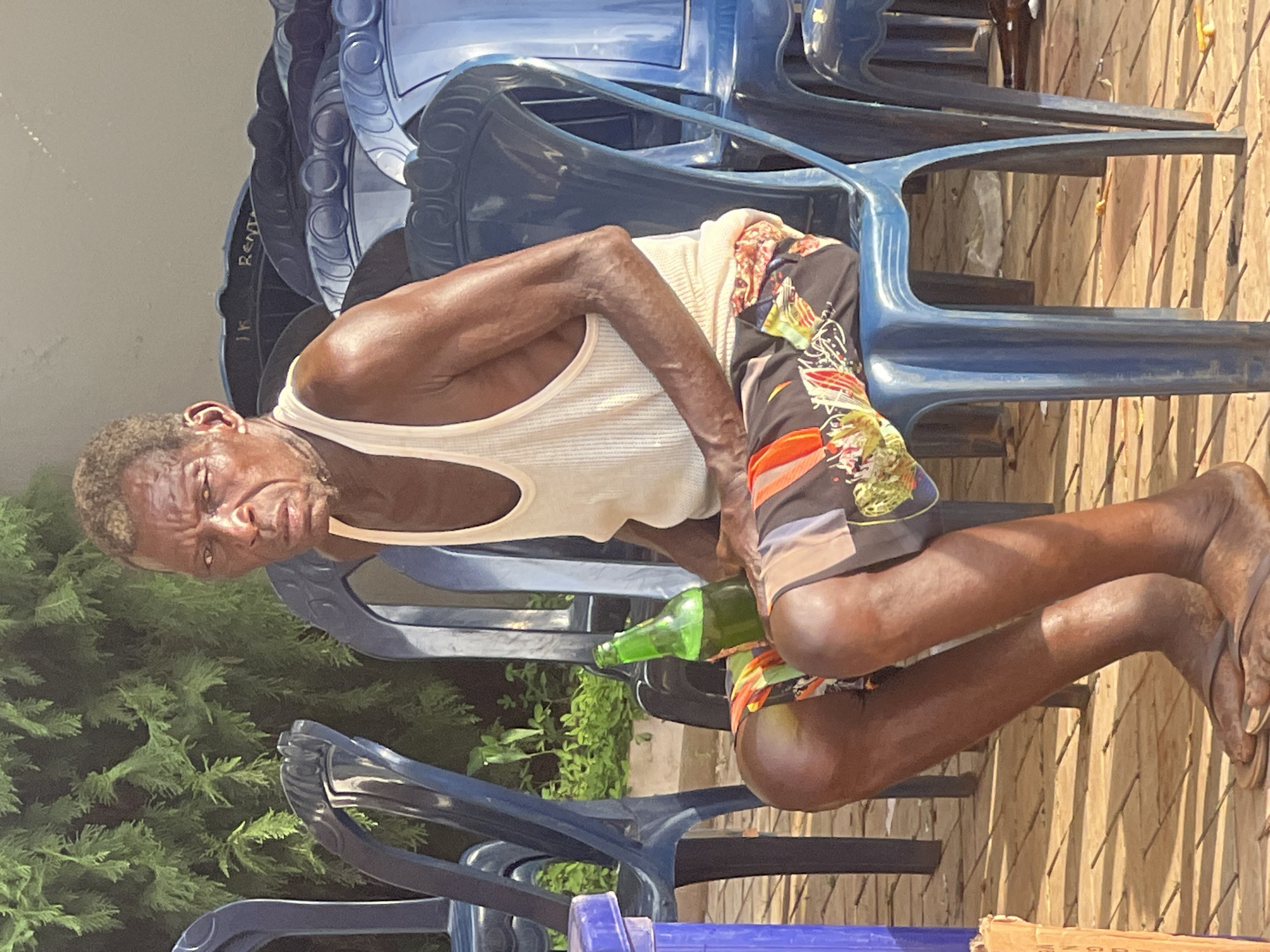 A man sits on a blue plastic chair in what appears to be an outdoor area. He is wearing a white tank top and patterned shorts. In his hand, he holds a green glass bottle. Several blue plastic chairs are stacked around him. The background includes greenery and a wooden fence. Given the context of Onitsha, Nigeria, the scene likely represents a casual, informal setting, possibly near a residence or small business.