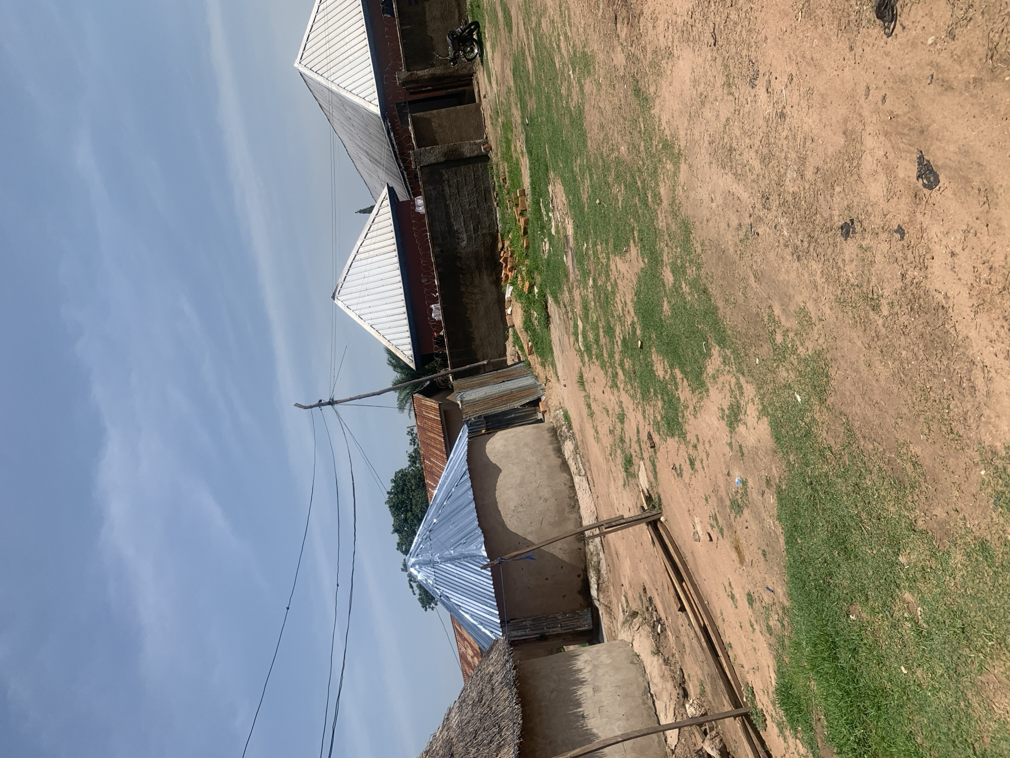 The image depicts a rural scene in Jauro Yinu, Nigeria, under a partly cloudy sky. The foreground is dominated by dry, reddish-brown earth with patches of sparse green grass. Several wooden planks are stacked in the middle ground.
To the left, a round, thatched-roof hut stands, made of mud or clay. Adjacent to it is another round mud structure with a corrugated metal roof. In the background, a long, low-lying building with a prominent, angular corrugated metal roof is visible. This building appears to be constructed with reddish brick or stucco walls and is topped with barbed wire fencing along its roofline. A concrete wall, also topped with barbed wire, extends from this building, enclosing a courtyard area. A parked motorcycle is visible near a doorway in this courtyard. Several piles of bricks are scattered in front of the concrete wall. Power lines are strung across the upper portion of the image, supported by a wooden utility pole. The overall impression is one of a semi-developed rural settlement.