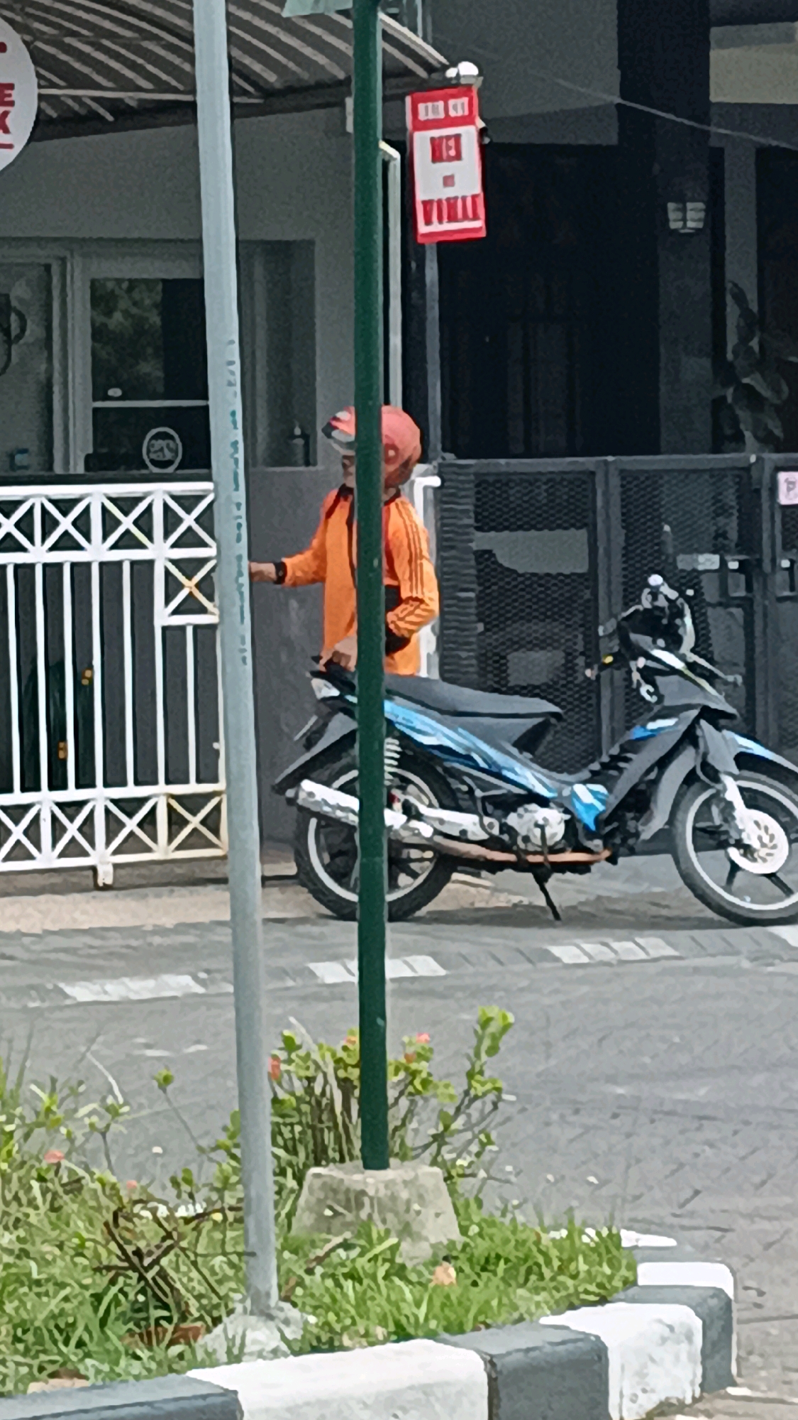 In Surabaya, Indonesia, a person wearing an orange jacket and an orange helmet is standing next to a black and blue motorcycle. The person is facing away from the camera and appears to be holding onto a white fence. The motorcycle is parked on a paved street. Behind the person and motorcycle, there is a dark-colored fence and a building. A red and white sign with text is hanging from a pole to the right of the person. In the foreground, there is a sidewalk with grass and some small plants.