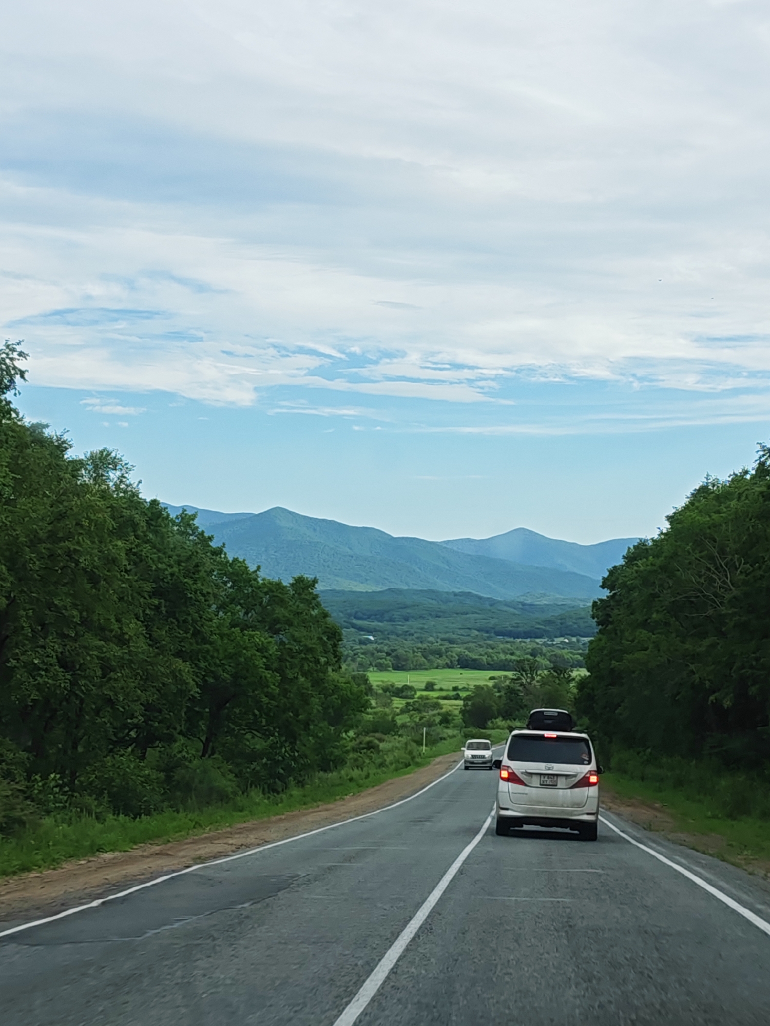 A slightly elevated, forward-facing, medium shot captures a two-lane asphalt road receding into the distance towards a lush green valley with rolling, blue mountains under a cloudy sky. The road is marked with white lines and has patches of wear. On the right side of the road, dense green trees line the shoulder, and on the left, a similar dense tree line borders the road. In the foreground, a white minivan with a roof carrier is in the right lane, and further ahead in the left lane is a white car. The sky above is a mix of blue and white clouds, with some clouds appearing wispy and others more textured. The overall scene suggests a scenic drive through a rural, mountainous region, likely in the city of Zolotaya Dolina, Russia, given the provided context of the location.