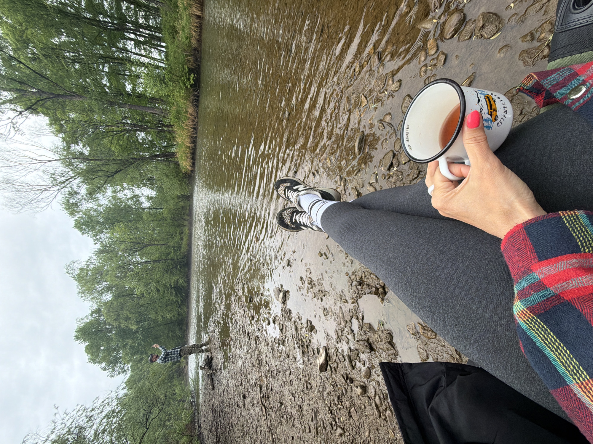 An eye-level, wide-angle shot shows a person sitting, likely on a bank, beside a body of water. Another person stands in the distance near the water, with trees lining the bank. The setting is likely rural.
The primary subject is a person from the knees up, seated with their legs slightly crossed. They wear grey leggings, leopard-print sneakers with striped white socks, and a red-and-blue plaid shirt. In their right hand, they hold a white enameled mug containing a brown liquid, possibly tea or coffee. Their fingernails are painted pink, and they wear a white ring on their ring finger.
To the left of the seated person, across a narrow, slow-moving body of water, another person stands on the rocky bank. This individual wears a plaid shirt and dark pants, and they appear to be waving or gesturing toward the camera.
The environment consists of a muddy and rocky bank transitioning into a water body, possibly a river or wide stream. The water appears murky. Along both banks are dense trees with green leaves, suggesting a forest or park. The sky is overcast, indicating a cloudy day. The location may be Trek, Russia.