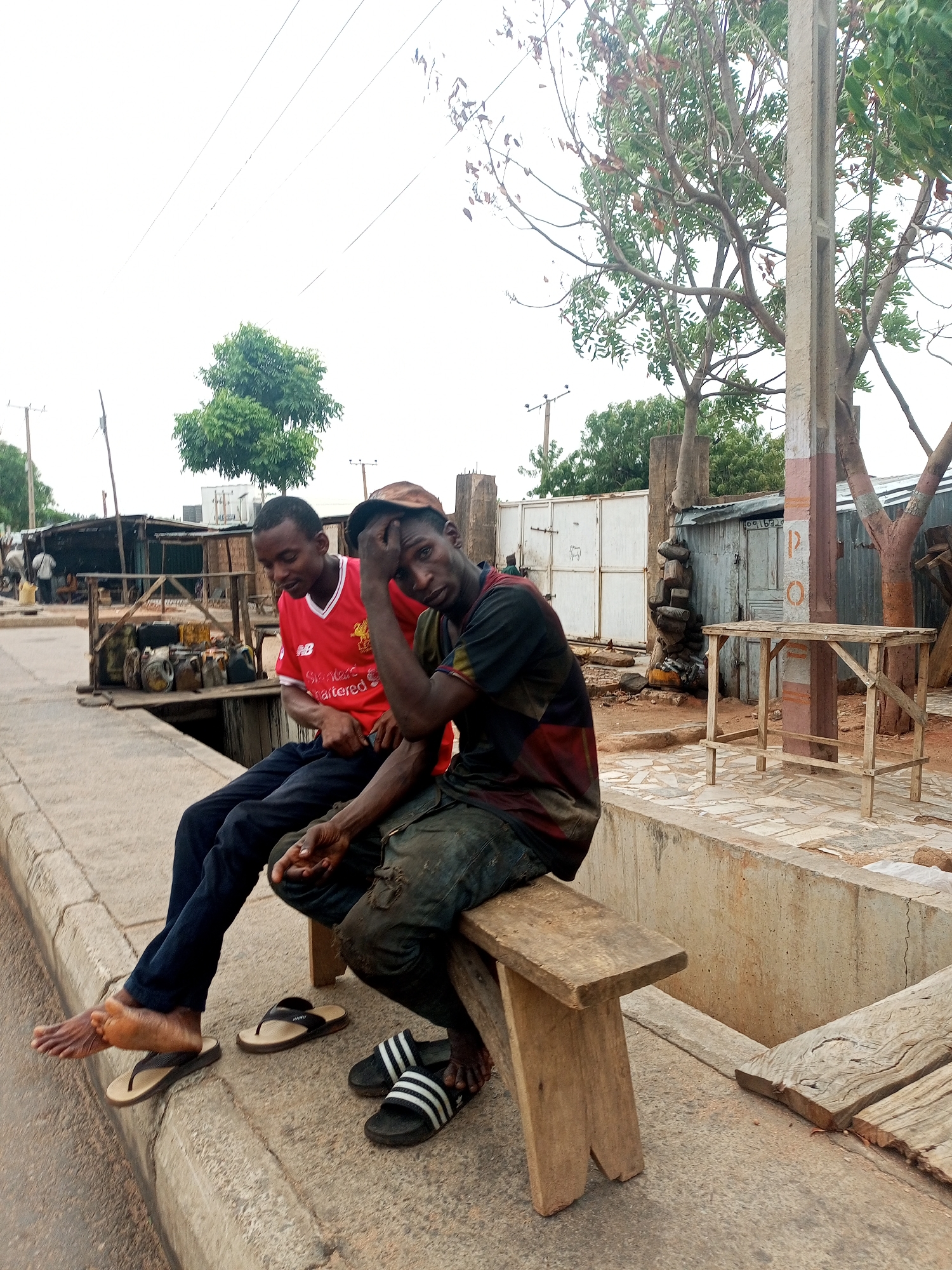 Two men are sitting on a wooden bench next to a street in Tempure, Nigeria. The man on the left is wearing a red Liverpool jersey and dark pants, with sandals next to his bare feet. The man on the right is wearing a dark patterned shirt, dirty pants, and Adidas sandals, with his hand on his head. Behind them, there is a street with buildings and trees. A utility pole is visible near the right side of the image.