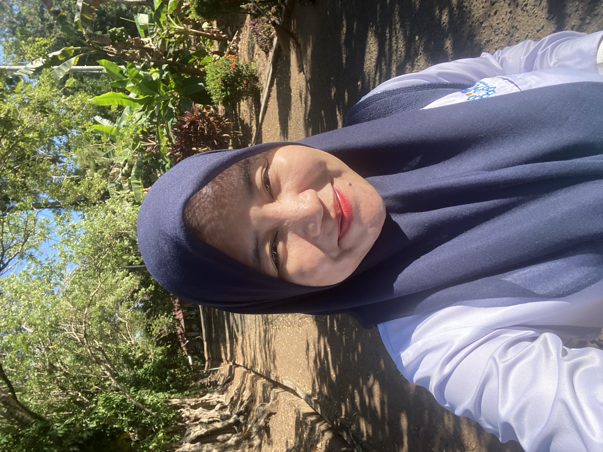 In Banjarbaru, Indonesia, a woman in a navy hijab and a white shirt takes a selfie outdoors. She is standing on a dirt path lined with trees on the left and lush vegetation on the right. The sun casts dappled shadows across the path. The woman is smiling and looking directly at the camera. A building with a red roof is visible in the distance. The background suggests a tropical or sub-tropical environment with abundant foliage.