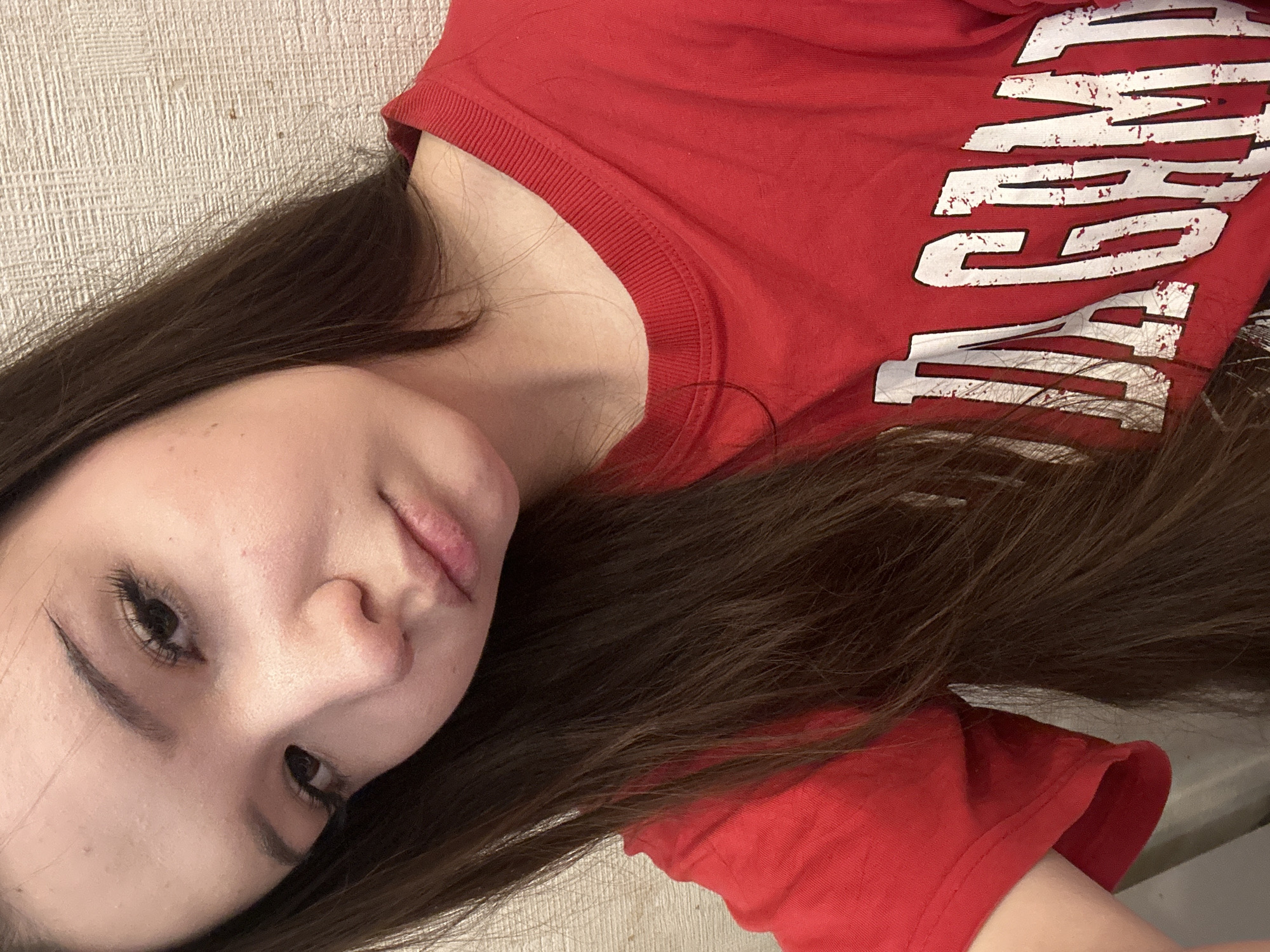 A young female is presented in a close-up, upper-torso portrait. Her head is tilted slightly to the viewer's left, with her gaze directed slightly downward and towards the camera. She possesses light skin, dark eyes, dark eyebrows, and prominent eyelashes. Her lips are slightly parted, showing some surface texture. Her long, dark brown hair is parted on her left side and flows over her shoulders and chest. She is dressed in a red, short-sleeved, crew-neck t-shirt with white, distressed block letters visible on the front, spelling 'JAGAM' with additional obscured characters to the right. The background consists of a light-colored wall with a repeating textured pattern. No specific geographical identifiers for Naberezhnye Chelny, Russia, or any other location, are discernible within the image.