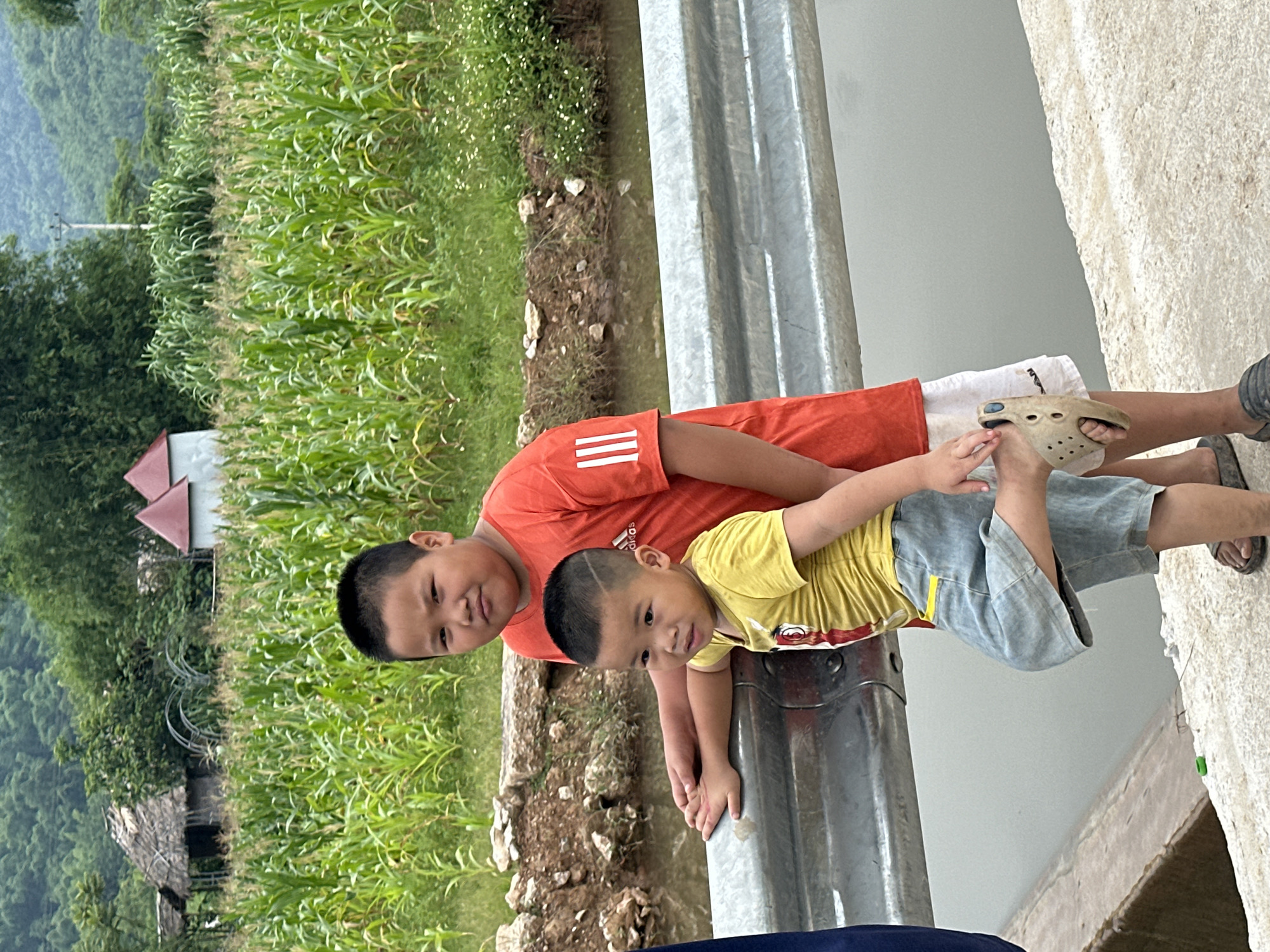 Two young boys, one older and one younger, stand in front of a cornfield in Mai Chau, Vietnam. The older boy, wearing an orange t-shirt with white stripes on the sleeve and white shorts, stands behind and to the right of the younger boy. The younger boy, wearing a yellow t-shirt and light blue shorts, leans against a silver metal barrier, with his left leg bent at the knee and his foot raised. Both boys have short black hair. In the background, a lush green cornfield stretches towards rolling hills covered in dense vegetation. A few simple structures, one with a thatched roof and another with red triangular roofs, are visible within the cornfield. The ground in the foreground is a textured, light-colored concrete or stone surface. The overall scene depicts a rural setting with elements of agriculture and simple village life.