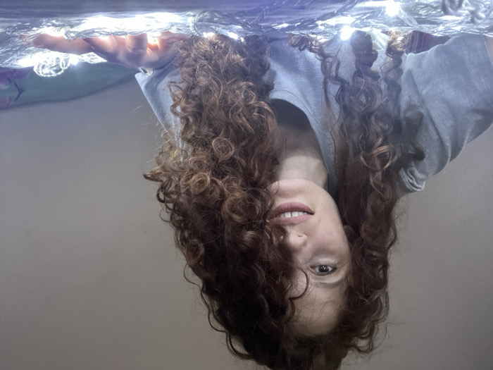 A female individual, appearing to be in her 20s to 30s, is depicted in a medium close-up, reclining in an indoor setting. She has long, voluminous, curly reddish-brown hair, fair skin, and a small stud piercing in her left nostril. She is wearing a plain grey short-sleeved t-shirt. The subject is looking directly at the camera with a subtle smile. Her right hand is visible in the lower right quadrant, resting amidst a dense entanglement of brightly illuminated white LED string lights. These lights are spread across a light-colored, soft surface, likely a bed or blanket, and serve as the primary light source, casting an upward glow. The background is a plain, light-colored, out-of-focus wall. No specific architectural or environmental markers are present within the image to determine its precise location within the city of Saint Petersburg, Russia.