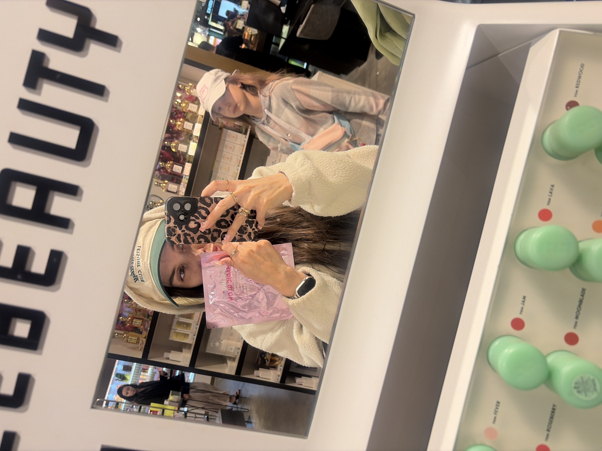 The image shows a person taking a mirror selfie in a retail store. The store appears to be a beauty or cosmetics shop, as evidenced by the shelves stocked with various products, including perfumes. In the foreground, the person is holding a pink packet and a smartphone with a leopard-print case. They are wearing a cream-colored fuzzy jacket and a tan baseball cap with green trim. Another person, possibly a child, is visible in the reflection of the mirror, wearing a plaid hoodie and a white baseball cap. In the lower part of the image, there is a display of several cylindrical green objects with labels, suggesting makeup or skincare products. The word "BEAUTY" is prominently displayed in large black letters on a white background above the mirror. There is no indication in the image that this scene is located in Saint Petersburg, Russia.