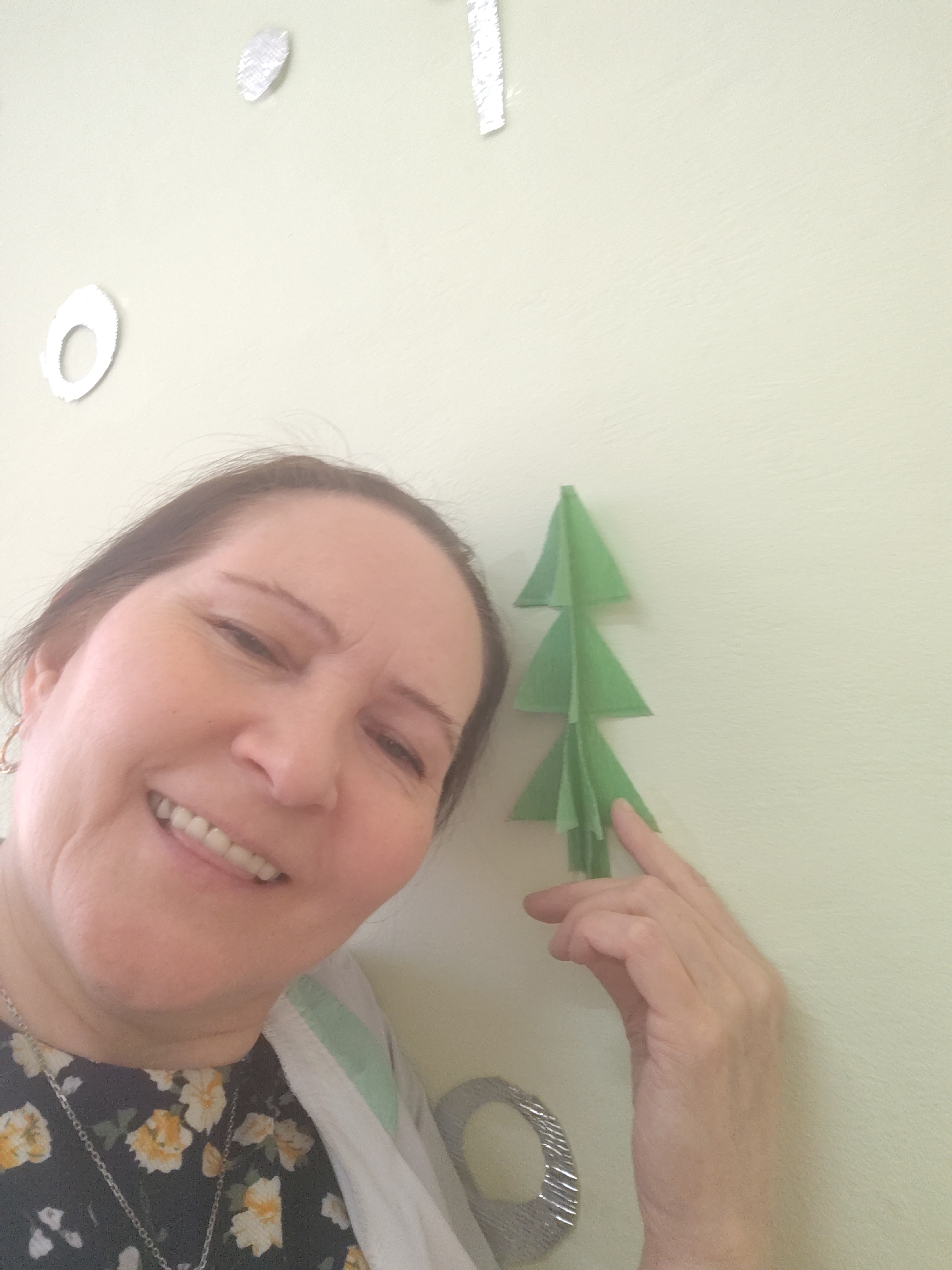 A smiling woman is captured in a close-up, slightly upward-angled shot, likely a selfie, within an indoor setting in Sarapul, Russia. She is positioned towards the left of the frame, looking directly at the camera with a wide, joyful smile that shows her teeth. She has medium-length, dark brown hair and is wearing a dark top with a yellow and white floral pattern, a silver chain necklace, and a light-colored cardigan with a subtle green stripe on the collar. A small, gold-colored hoop earring is visible in her left ear.
To her right, on a light greenish-beige wall, is a decorative, three-dimensional green paper Christmas tree. Her right hand is visible, with her index finger gently touching or pointing to the lower part of the paper tree. The wall also features several metallic, silver-toned decorations, including a circular outline, a leaf-like shape, and a textured rectangular strip, suggesting a festive or personalized space. Another large, textured silver ring is partially visible below her right arm. The overall lighting is bright and even, indicating daytime. No text is visible in the image.