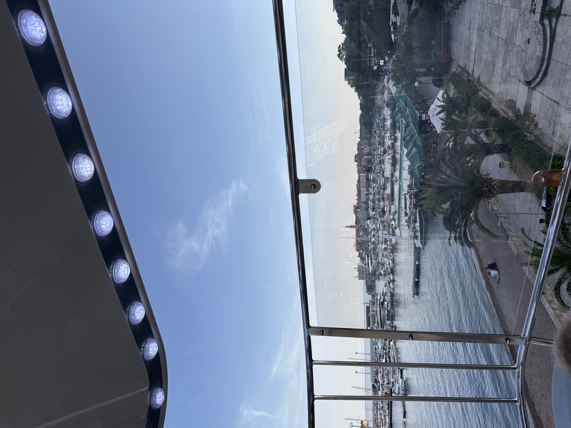 The image captures a scenic coastal view of Budva, Montenegro, from an elevated perspective, possibly from a high balcony or a cabin on a Ferris wheel. The original image is rotated 90 degrees clockwise, so the description assumes it has been virtually reoriented.
In the foreground, the top part of a structure is visible, featuring a row of round, illuminated lights embedded in a gray surface, suggesting artificial lighting for a canopy or viewing platform. Below this, a clear glass barrier or railing with metallic supports extends across the frame, offering an unobstructed panoramic vista. Reflections of the sky and subtle clouds can be seen on the glass.
The background reveals a vibrant marina bustling with numerous boats, including yachts and smaller vessels, docked along a calm body of water. A paved promenade runs parallel to the waterfront, lined with green awnings of what appear to be cafes or restaurants. Several lush palm trees are scattered along the promenade and a small sandy beach. A few individuals can be seen relaxing on the beach, and others are walking along the promenade. Beyond the immediate coastline, a dense cluster of buildings with varying architectural styles, including some with reddish roofs, rises up a hillside. Further back, verdant hills covered in trees provide a natural backdrop to the urban and maritime scene.
The time of day appears to be daytime, likely late afternoon, under a clear blue sky with sparse, wispy clouds, indicating pleasant weather. No discernible text is visible within the image. The scene conveys a peaceful and picturesque atmosphere of a popular Mediterranean coastal town.