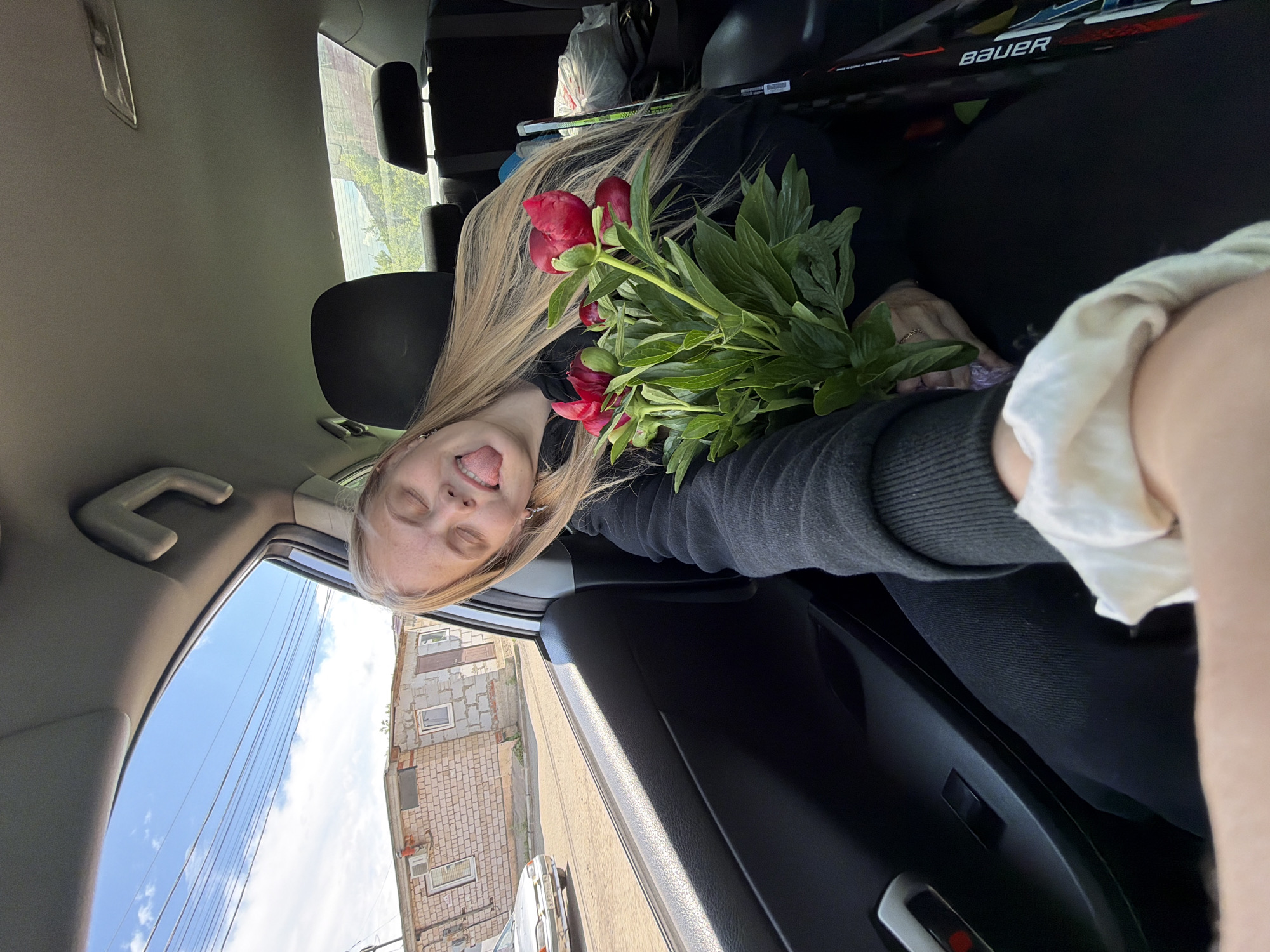 A woman is lying in a car, holding a bouquet of red peonies. She is sticking her tongue out with her eyes closed. She has light blond hair and is wearing a black top. In the background, there is a Bauer hockey stick. Through the car window, a view of a city building in Chaikovsky, Russia, is visible.