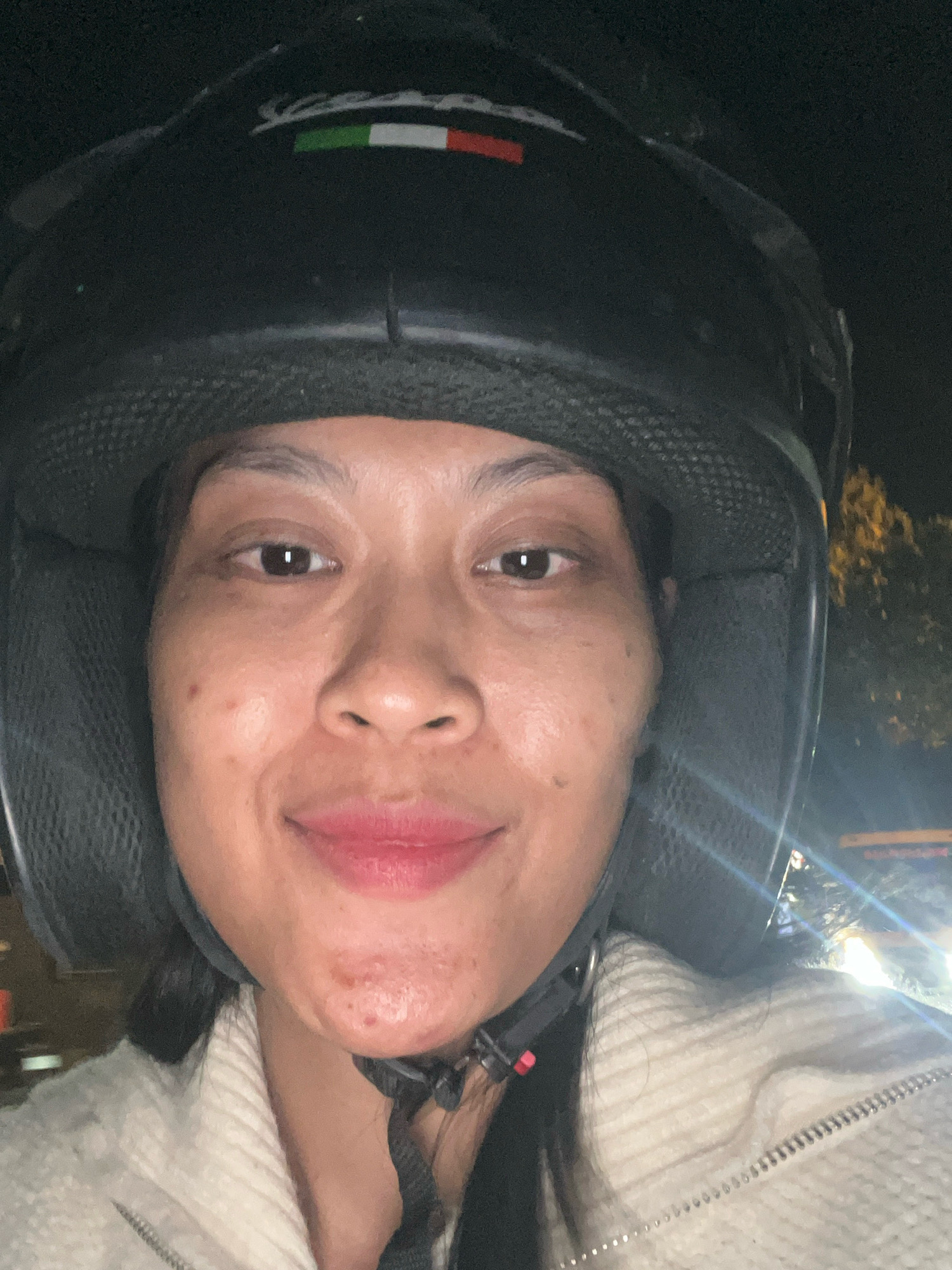 A close-up, front-facing selfie shows a woman wearing a black helmet with a Vespa logo on the front, featuring the Italian flag colors. She has medium-toned skin with some blemishes on her cheeks and chin. Her lips are painted with a reddish-pink lipstick. She is wearing a light-colored, textured jacket. The background is dark, suggesting nighttime, with blurred lights and foliage visible, hinting at an outdoor urban environment, possibly on a street in Kecamatan Pati, Indonesia.