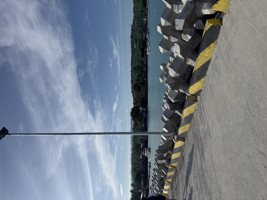 This image depicts a pier or dockside area in Toyapakeh, Indonesia, under clear daylight conditions. The foreground consists of a grey concrete pier surface with visible textures and shadows. Along the right edge of the pier, a series of interlocking, multi-faceted grey concrete tetrapods form a barrier or breakwater. These tetrapods feature diagonal yellow and black striped paint at their base. Approximately in the center of the image, a tall, slender metallic pole, likely a lamppost, extends vertically from the pier towards the sky, topped with a dark, rectangular fixture.
To the far left, a partial view of an individual, dressed in dark clothing, is visible, walking away from the camera. Beyond the pier and tetrapods lies a body of turquoise-blue water, indicative of a strait or bay. Several boats are present on the water; a white motorboat with a cabin is visible in the mid-distance, moving from left to right. Other smaller vessels are moored or scattered closer to the distant shoreline. The background features a lush, green, and hilly coastline covered with dense vegetation and trees. The sky above is a bright blue with scattered, wispy white clouds. The overall scene suggests a coastal port or transit point.